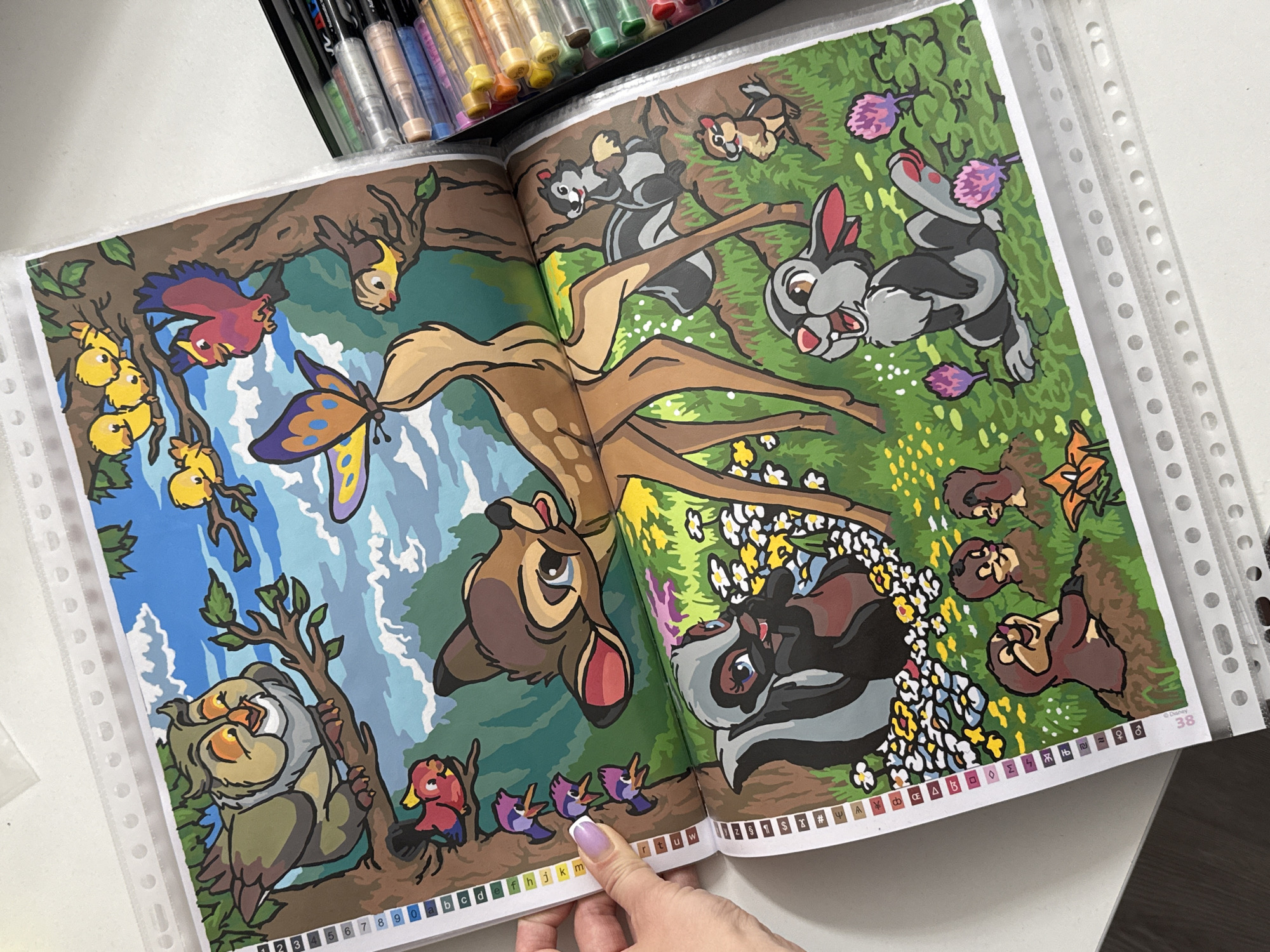 The image shows a close-up of a paint-by-numbers coloring book featuring characters from Disney's Bambi. The book is open to a page with a vibrant illustration of Bambi, Thumper, Flower, and other forest creatures. The style of the illustration is cartoonish and colorful, with clear outlines and large areas of flat color. The image is well-lit, and the colors of the artwork are bright and saturated. A box of colored markers or pens is visible to the right of the book, suggesting that someone is in the process of coloring the picture. The book is placed on a white surface, likely a table. There are no identifiable landmarks or elements that would indicate a specific location within the city of Łódź, Poland.