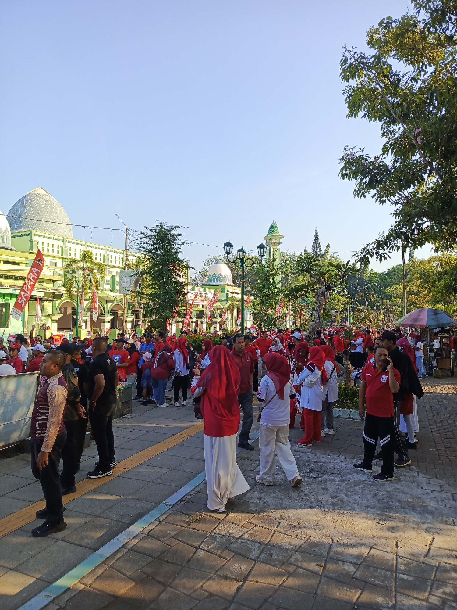 The image depicts a gathering of people in front of a mosque, likely in the city of Semampir, Indonesia. The mosque features prominent green domes and white walls, with decorative green accents on its facade and minarets. Many individuals in the crowd are wearing red attire, suggesting a coordinated event or celebration. Red and white flags, some bearing the word "SAFARA," are visible throughout the scene, further indicating a festive or patriotic occasion. The ground is paved with interlocking stones, and there are trees and streetlights present, suggesting an outdoor public space. Some people are standing, others appear to be walking or conversing. The overall atmosphere seems to be that of a community event taking place during daylight hours.