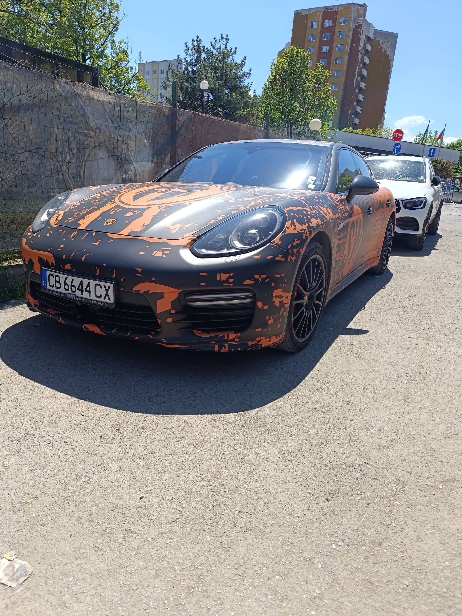 The image shows a black and orange Porsche Panamera parked on an asphalt street in Sofia, Bulgaria. The car has a Bulgarian license plate that reads "CB 6644 CX". Next to the Porsche, there is a white Mercedes SUV. In the background, there is a weathered building with a "STOP" sign visible. A fence is seen behind the Porsche. The weather appears sunny and bright.