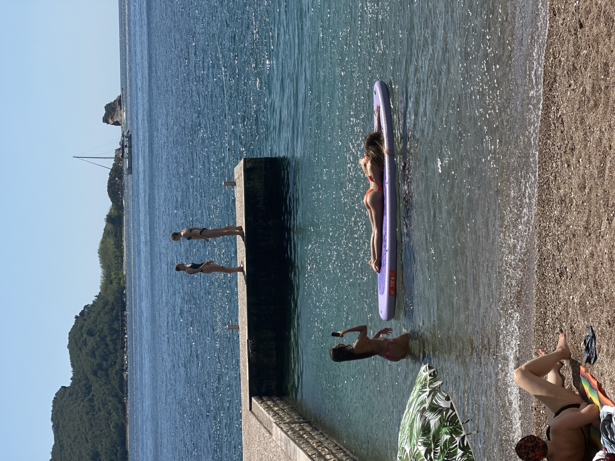 The image depicts a sun-drenched scene at a beach in Budva, Montenegro. In the foreground, a person wearing a black bikini and a patterned headscarf is lying on a colorful towel on the pebbly beach, facing away from the camera. Adjacent to them are a pair of dark flip-flops.
In the clear, shallow water, a young woman in a pink bikini is standing, holding a smartphone and appearing to take a photo. Further out in the water, another woman in an orange bikini is lying on a purple inflatable paddleboard, with her body stretched out and her face turned away from the camera.
A stone pier extends into the blue sea, with two young women in bikinis standing on its edge. One is wearing a black bikini, and the other is wearing a dark bikini. In the background, a sailboat with a tall mast is visible on the water, with a rocky island formation in the distance. Beyond the water, a lush, green, tree-covered hill rises, with buildings nestled at its base. The sky is clear and blue.