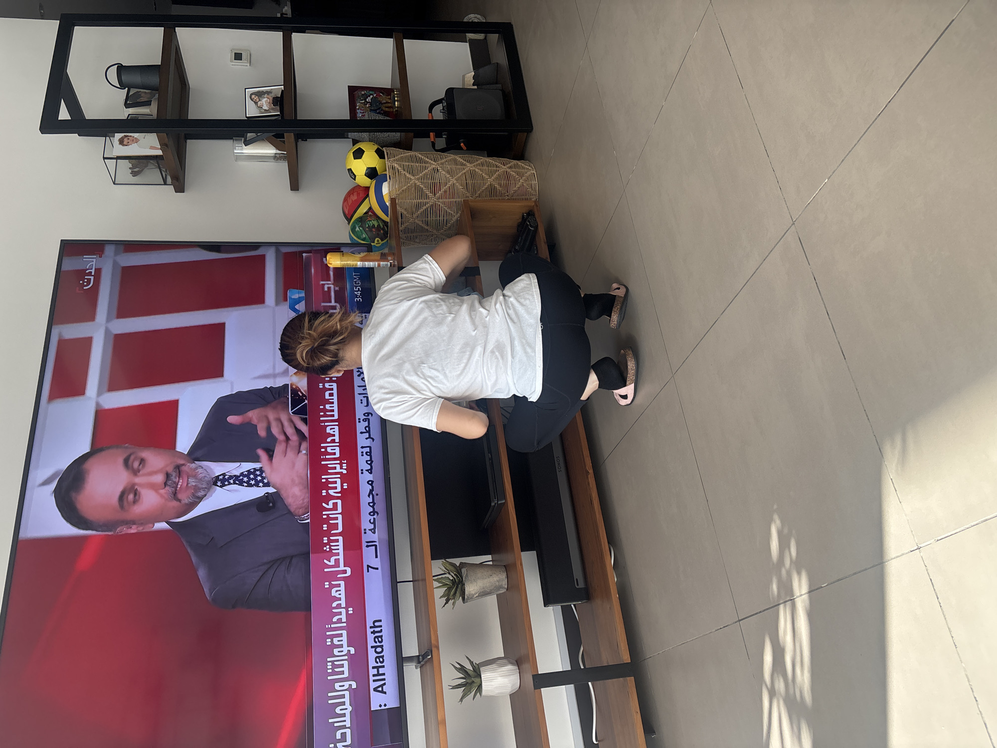 In a modern apartment in Dubai, United Arab Emirates, a woman is crouched on the floor in front of a large television. The TV displays a news program with an anchor wearing a suit and tie. The woman, with her hair tied back in a bun, wears a white t-shirt and black leggings, with pink sandals. She appears to be interacting with the electronics below the TV, possibly a soundbar or game console.
The living room features a wooden media console with several shelves. On the shelves are decorative plants, a soundbar, and other electronic devices. To the right of the television, a tall, black metal shelving unit holds various decorative items, including framed photos and a woven basket filled with sports balls. Sunlight streams in from the left, casting shadows on the tiled floor. The overall scene suggests a casual, domestic setting in a well-appointed urban dwelling.