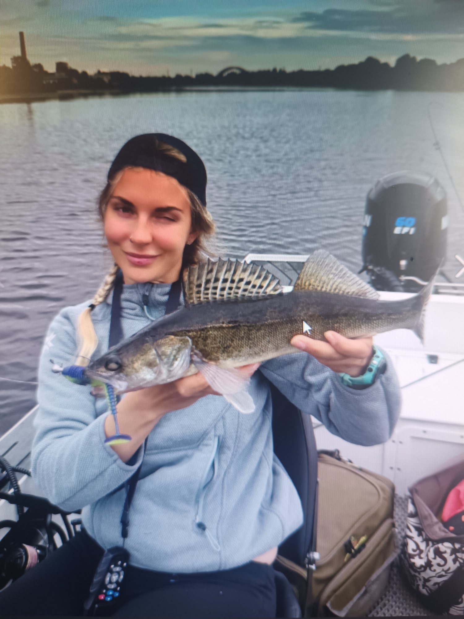 A young woman wearing a black baseball cap and a light blue hooded sweatshirt is holding a caught fish in her hands. The fish is long and slender with a dark patterned back and a lighter belly. The woman is on a boat, with a body of water behind her and a boat motor visible to her right. In the distance, the shoreline of a city is visible with buildings and trees. The sky is overcast, suggesting it may be evening or a cloudy day. The overall scene depicts a successful fishing trip. The image does not provide any information about the location being the city of Klyuchi, Russia.