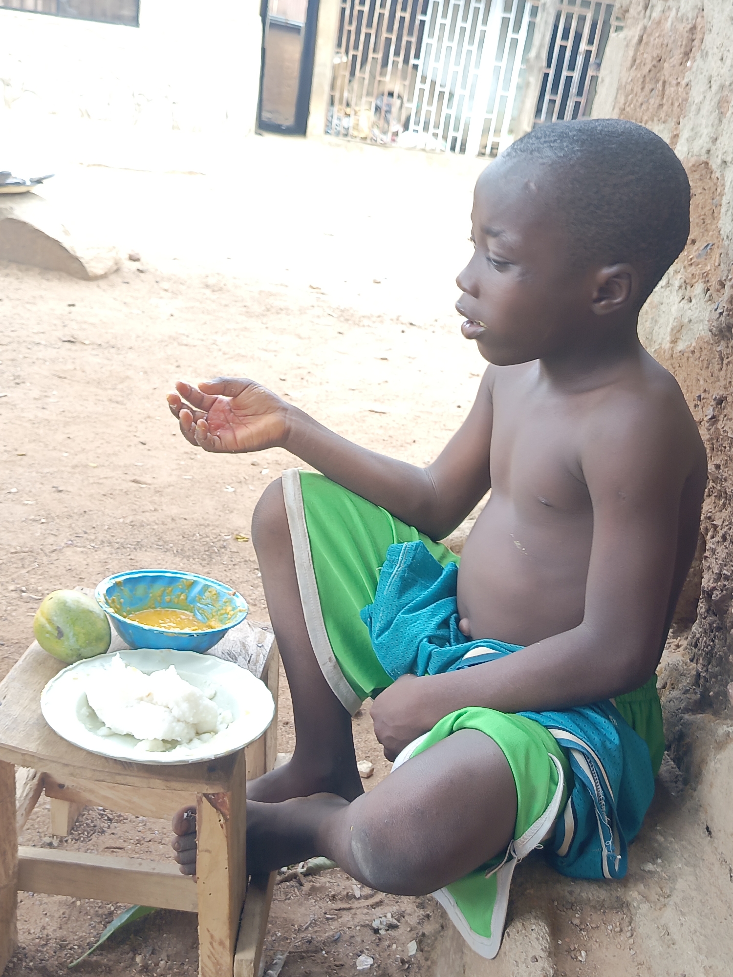 A young boy with dark skin and short dark hair is captured in a side profile, seated on the dirt ground next to a rough, textured wall. He is shirtless, wearing green athletic shorts over what appears to be blue mesh shorts. His right hand is slightly cupped, palm up, suggesting he is preparing to eat or has just eaten.
In front of him, on a small, simple wooden stool, is a meal. A white plate holds a portion of a white, starchy food, likely fufu. Beside the plate, a vibrant blue bowl contains a yellow, chunky stew or soup. A whole, unripe green mango rests next to the food items on the stool.
The setting appears to be outdoors in natural daylight. The ground is dusty and earthen. In the background, there's a light-colored building with a dark-framed opening and metal bars, possibly a window or gate. The overall scene conveys a simple, everyday moment in a likely rural or semi-urban environment, consistent with Akwanga, Nigeria. The boy's expression is calm and focused on his hand and the food. There is no visible text in the image.