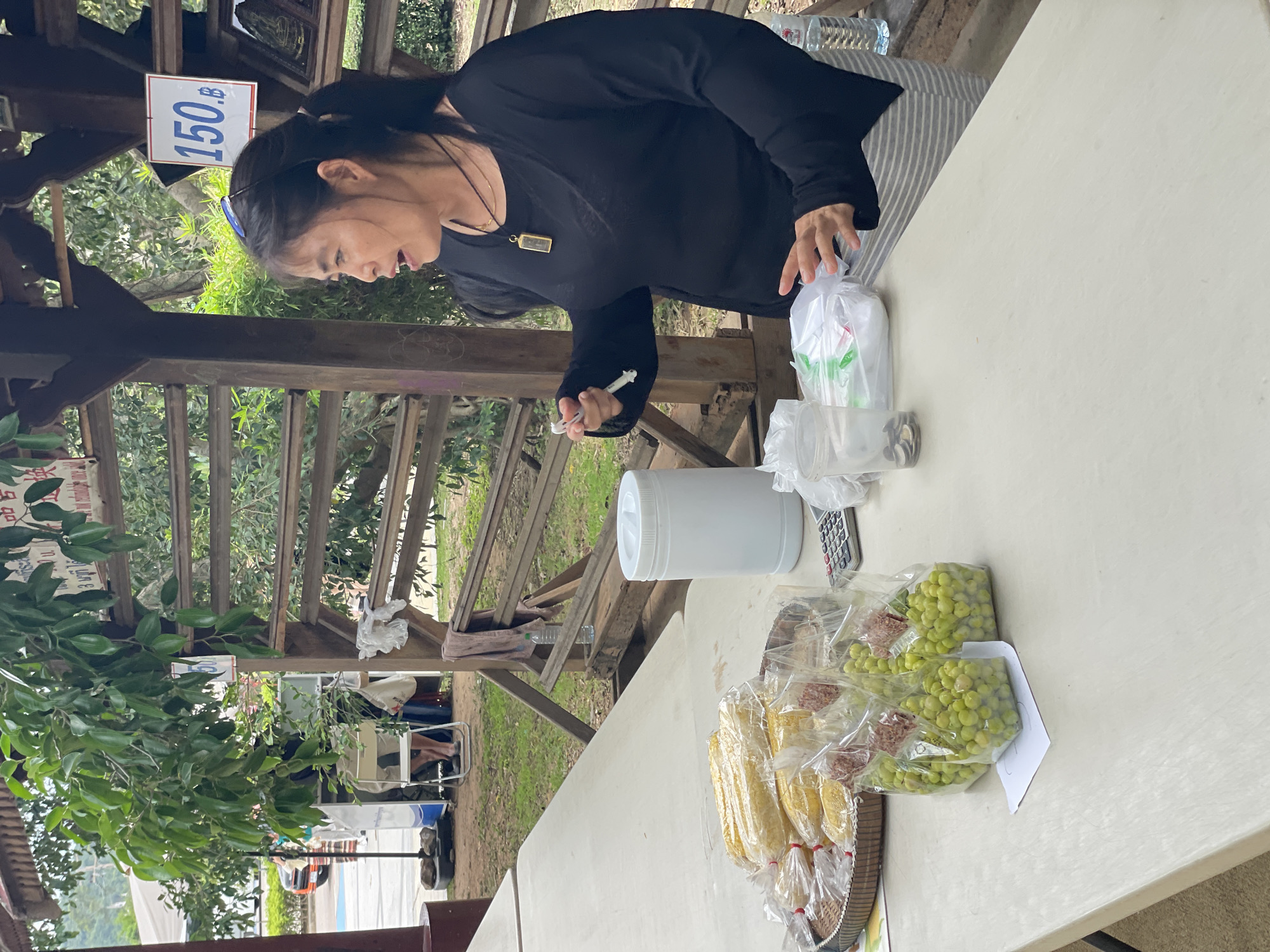 A medium shot captures a woman standing behind a table laden with food items in Na Chom Thian, Thailand. She is wearing a black long-sleeved shirt and striped pants. Her hair is tied in a ponytail, and she is looking to her left, with her mouth slightly open as if speaking. She is holding a small white object in her right hand.
On the table, in front of her, are several plastic bags filled with corn and green peas. A woven basket holding more corn is visible to the left. To the right of the woman, a white cylindrical container, a calculator, and a clear plastic cup containing coins are present. A water bottle stands to the far right.
Behind the table, a wooden structure with horizontal planks is visible, and a sign displaying "150 B" is mounted on it. Green foliage forms the background. The overall scene suggests a market stall or an outdoor food vendor.