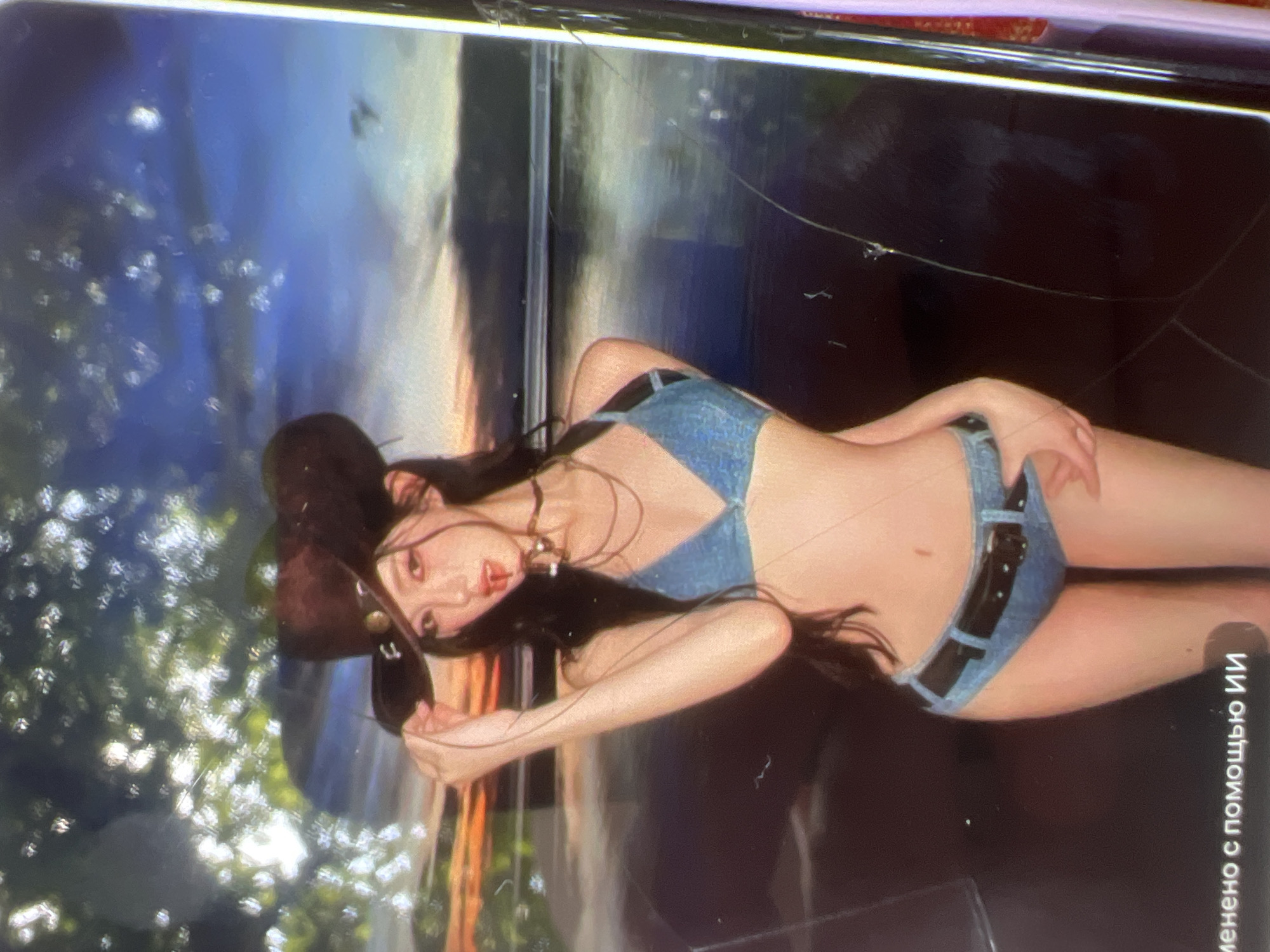 The image shows a young woman wearing a cowboy hat, a denim bikini top, and denim shorts with a black belt. She is holding the brim of her hat with her right hand and her left arm is bent at the elbow. She has long dark hair and is wearing a choker necklace. The background appears to be a sunset over water, with some foliage in the upper left corner. There are some scratches and reflections on the surface of the image. The text at the bottom left corner reads "именено с помощью ИИ", which translates to "modified with AI". There is no discernible information in the image that indicates a location in the city of Kara-Dıykan, Kyrgyzstan.