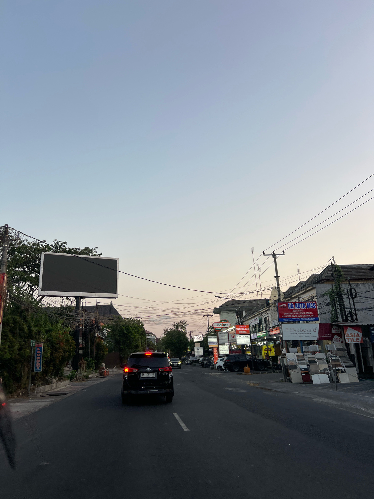 The image shows a street scene in Kuta, Indonesia, during the late afternoon or early evening. The sky is a gradient of pale blue to soft orange, indicating the setting sun. A black SUV is driving away from the camera on the right side of the road. Several commercial establishments are visible along the street, with illuminated signs in various languages. One prominent sign reads "UD. KUTA MAS" and advertises sanitary ware, ceramics, and bathroom accessories. Another sign for "ind@bat" is also visible. There are several power lines and utility poles overhead. In the foreground, on the left side of the road, a large, dark digital billboard is mounted on a pole. The street is paved and appears to be a busy thoroughfare with some parked vehicles. Trees and foliage line the left side of the road. The overall impression is of a commercial street in a tropical urban environment.
