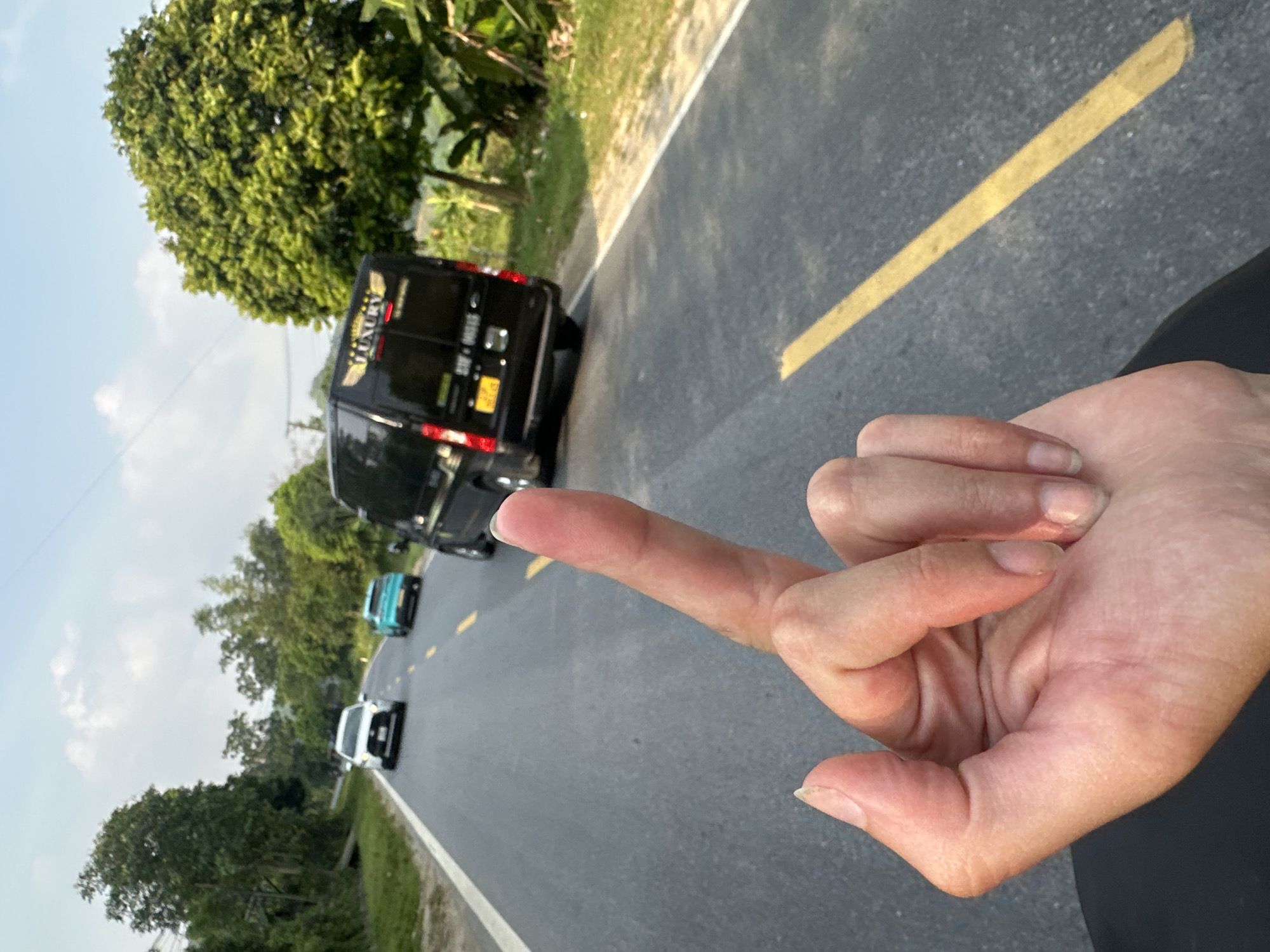 The image presents a tilted, eye-level view, focusing on a human hand in the immediate foreground, with the middle finger extended outward. This gesture appears directed towards a road scene in the background, conveying defiance or insult.
The setting is an asphalt road in Vị Xuyên Commune, Vietnam, clearly marked with a solid yellow line down its center and a white line along its right shoulder. Several vehicles are visible on the road. Most prominent is a black passenger van, possibly a commercial vehicle, with the word "LUXURY" clearly visible on its rear. Below "LUXURY", a Vietnamese license plate appears to display "30A-000.35". Further down the road, a turquoise car and a white car can be seen.
The road is bordered by dense, lush green trees and tropical foliage on both sides, indicative of a verdant rural or semi-rural landscape. The sky above is clear with scattered clouds, suggesting a bright daytime, likely late morning or early afternoon, with ample natural light.