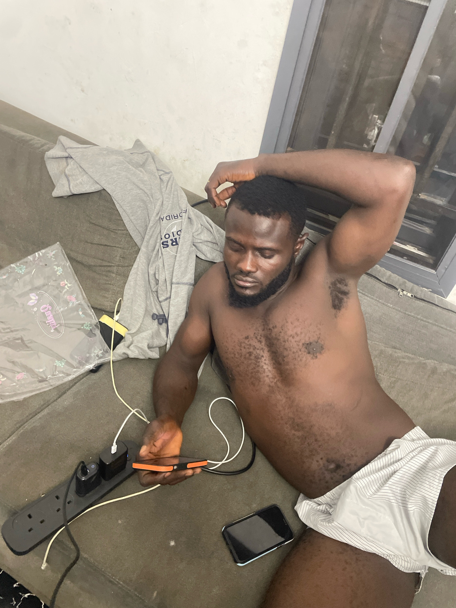 A Black man with a beard and short hair is lying on a couch in an indoor setting, possibly an apartment in Abuja, Nigeria. He is shirtless and wearing striped shorts. One arm is raised behind his head, and his eyes are closed. Next to him on the couch are a grey t-shirt, a plastic-wrapped item with a floral pattern, and a black power strip with several devices plugged into it, including two phone chargers. A smartphone is held in his right hand, connected to one of the chargers by a white cable. Another smartphone is visible on the couch to his lower right. The couch appears to be upholstered in a dark, textured fabric. A window with grey frames and what looks like a security grate is visible in the background. The lighting suggests it is daytime, with ambient light coming from an unseen source.