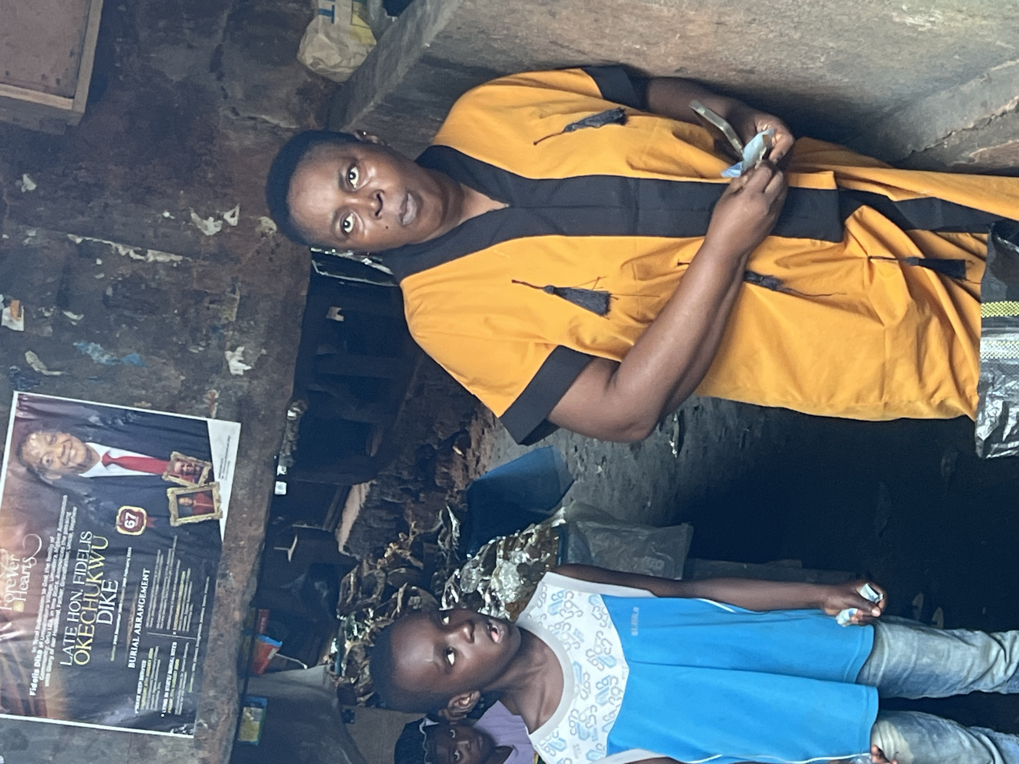 The image captures a scene in Nkpor, Nigeria, focusing on three individuals. In the foreground, a young boy wearing a blue and white sleeveless shirt and denim jeans looks to his left with his mouth open, his eyes wide. To his right stands a woman in an orange and black patterned garment, her gaze directed upwards. Her hands are clasped, and she appears to be holding a few folded bills. In the background, a younger child in a purple shirt and dark head covering is partially visible. Behind the people, a poster with a man's photograph and text announcing funeral arrangements for "Late Hon. Fidelis Okechukwu Dike" is prominently displayed. The environment appears to be a dimly lit, possibly open-air market or a similar outdoor space, with weathered walls and debris scattered around.
