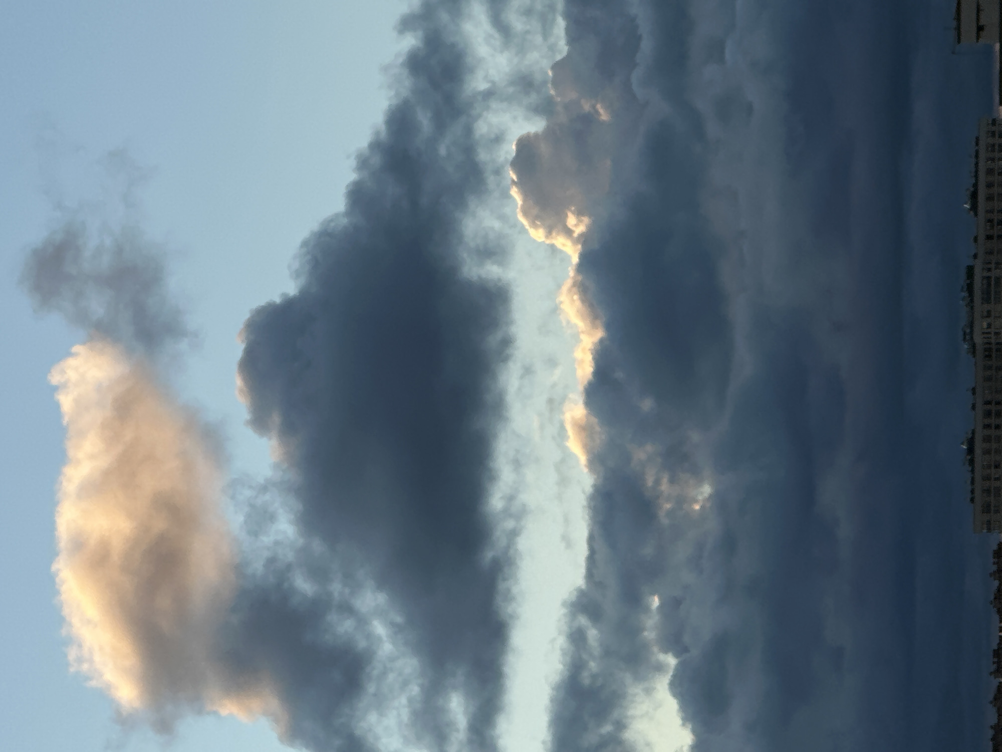 The image displays a dramatic sky filled with dark, heavy clouds, illuminated by a soft, warm light from the setting or rising sun peeking through. The upper portion of the sky features lighter, wispy clouds with a golden hue, contrasting with the predominantly dark, stormy-looking clouds that dominate the middle and lower sections. Below the clouds, a city skyline is visible, characterized by the silhouettes of buildings. Specifically, the architecture suggests apartment blocks, indicating an urban environment. The overall atmosphere is one of impending weather change, with the vibrant sky contrasting against the subdued urban landscape. The location is specified as Dnp Ryzhiki, Russia.