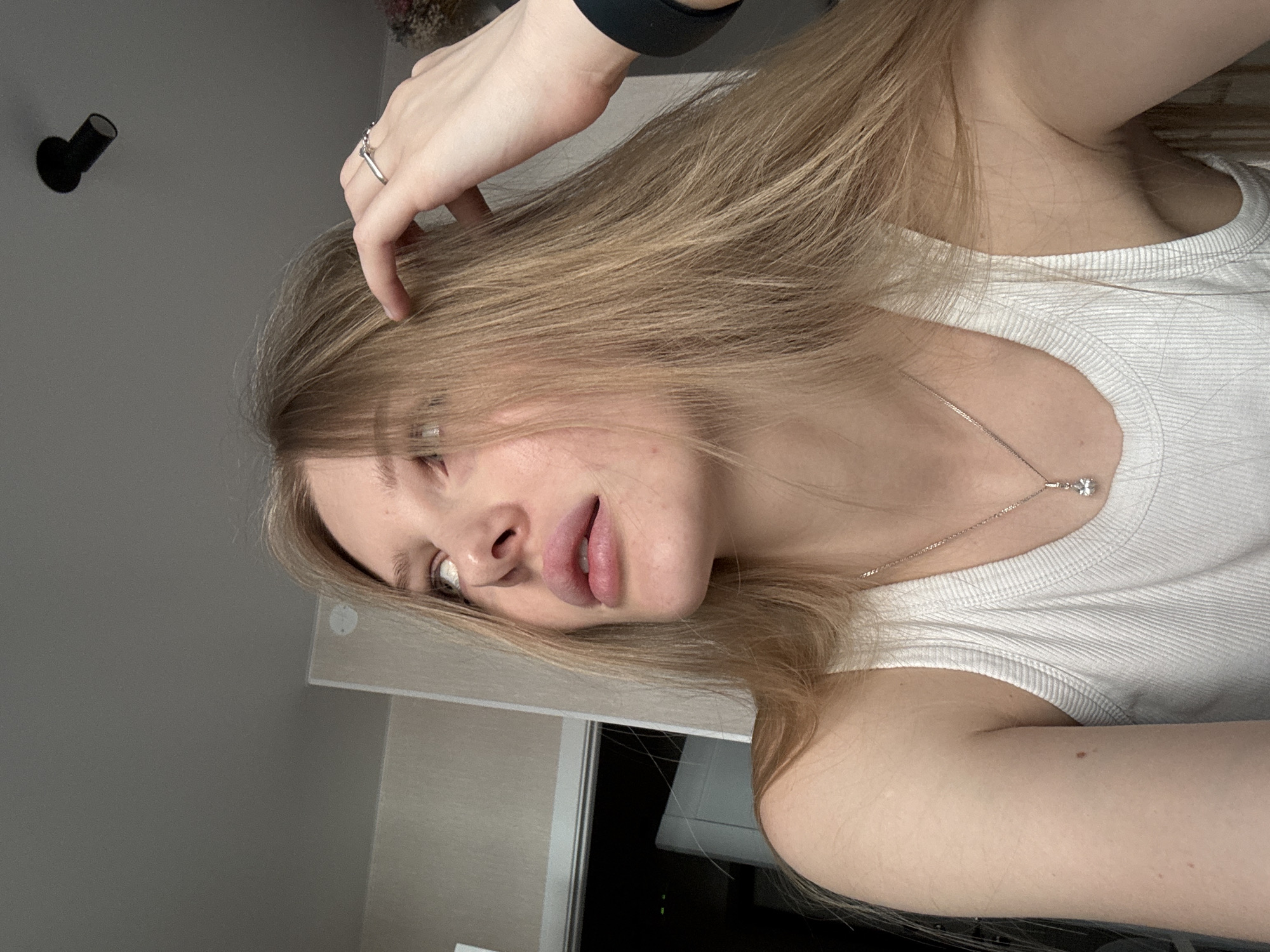 A young woman is pictured indoors. The image is oriented at an oblique angle. The woman has light skin, blonde hair and is wearing a white ribbed tank top and a silver necklace with a flower pendant. She has a ring on one of her fingers and a dark band on her wrist. Her hand is resting on her head, ruffling her hair. A black light fixture is visible on the wall in the upper left corner of the frame. A light gray wall and a combination of gray and dark surfaces appear in the background. Based on the visual information, there is no information that can confirm the location in Perm, Russia.