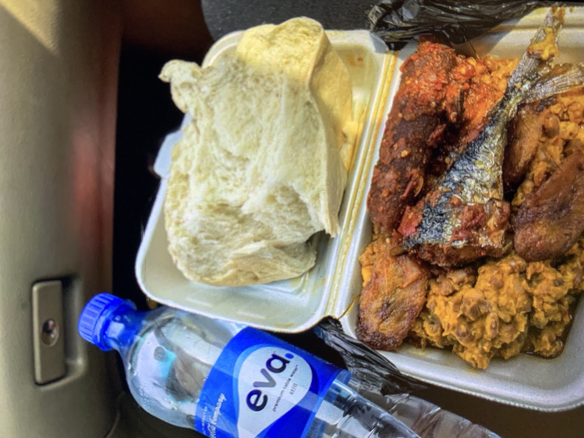 A still life depicts a meal and a bottle of water, likely photographed indoors. The meal is presented in a white styrofoam container separated into two sections. One section holds several pieces of what appears to be pounded yam or fufu, while the other compartment contains an assortment of food, including fried fish with its tail fin visible, fried plantains, and a stew of beans. A bottle of "eva" brand premium table water is placed next to the container. The background features part of a car's interior. The city of Rafin Par, Nigeria is not recognizable in this image.