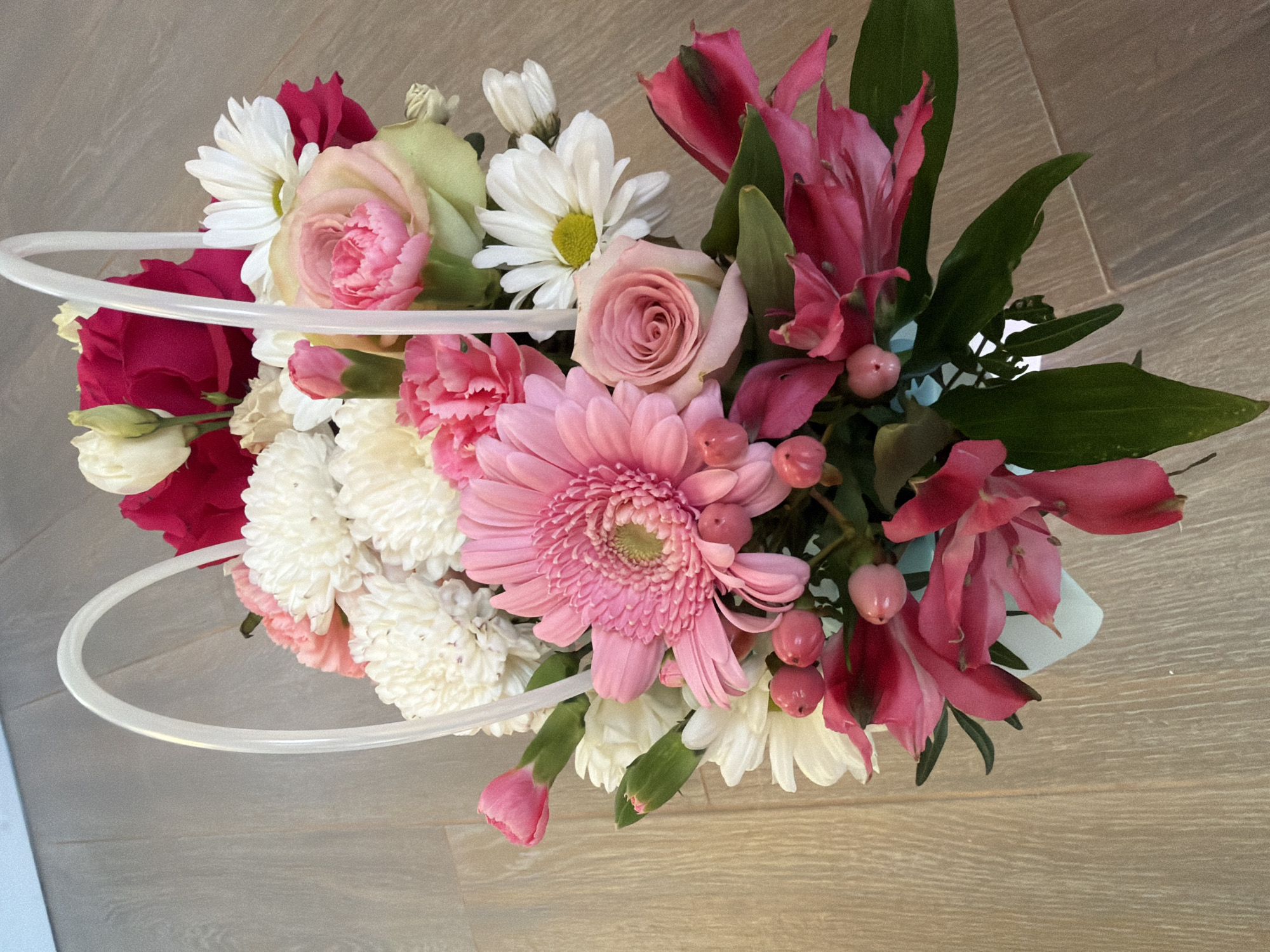 A high-angle shot shows a bouquet of flowers in a white basket with a clear plastic handle, placed on a light-colored wooden floor. The bouquet contains various flowers in shades of pink, white, and dark red, including roses, gerberas, carnations, daisies, and alstroemerias, as well as small pink berries. The background is a blurred light blue wall. The setting is indoors in the city of Uzyukovo, Russia.