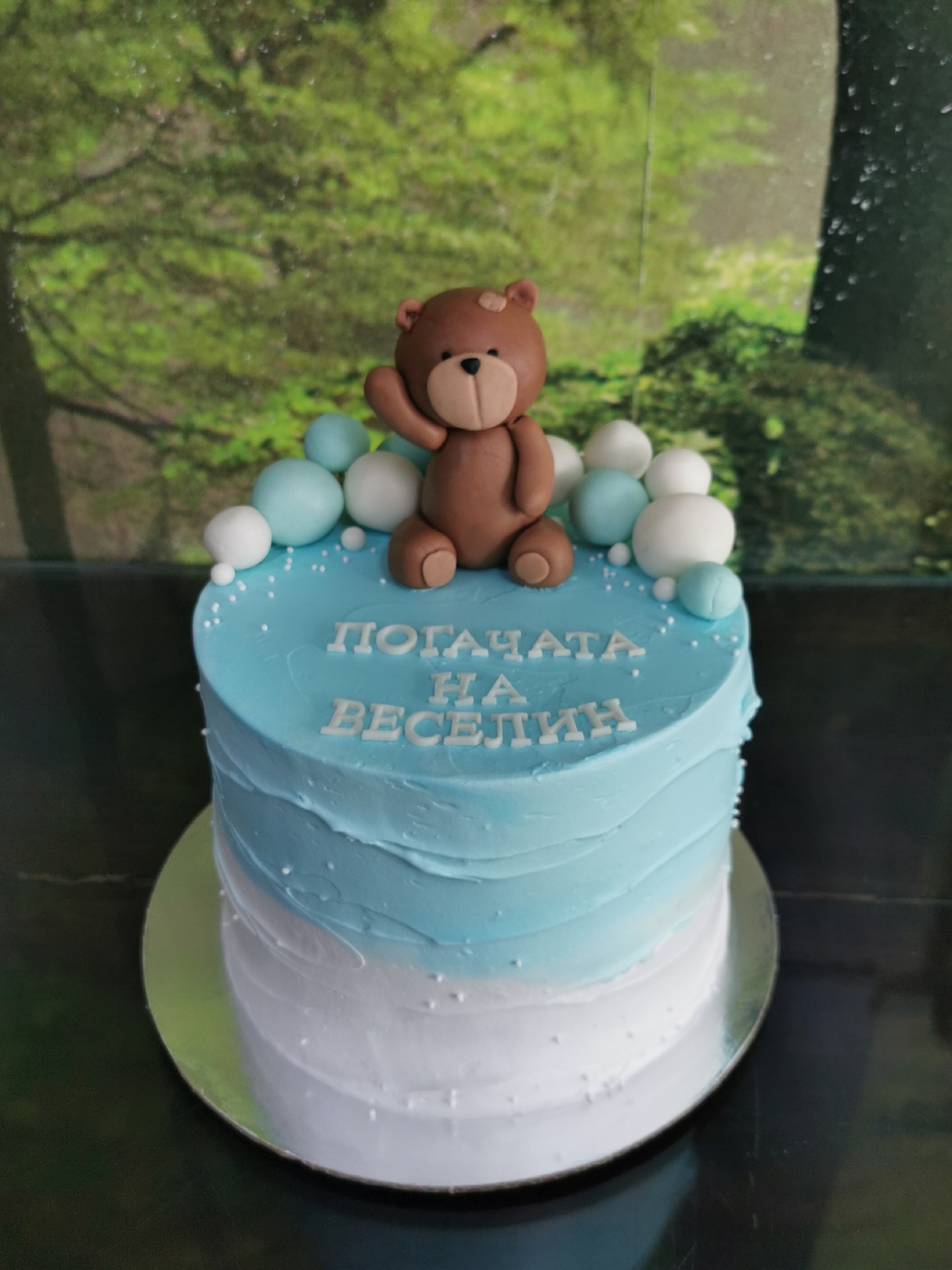 This media file displays a celebratory cake, likely for a baby boy, against a blurred green backdrop resembling foliage. The cake is round and features two colors of frosting: white on the bottom half, smoothly transitioning to a light blue on the top half, with a textured, swirled finish. Small white sugar sprinkles are scattered across the blue frosting.
A prominent decoration on top of the cake is a brown teddy bear figurine, with lighter brown snout and inner ears, sitting and appearing to wave. Around the teddy bear, and along the top edge of the cake, are spherical sugar paste decorations in varying sizes, in both light blue and white.
The top surface of the cake features white block letters in Bulgarian, spelling out "ПОГАЧАТА НА ВЕСЕЛИН" (POGACHATA NA VESELIN), which translates to "VESELIN's POGACHA". A pogacha is a traditional bread, often made for special occasions, and "Pogacha" is also a term used for the baby welcome ceremony in Bulgaria.
The cake rests on a reflective silver cake board, which is placed on a dark, possibly wooden, surface. The lighting is even, suggesting an indoor setting. Given the text and cultural context, the cake is prepared for a celebration in Varna, Bulgaria.