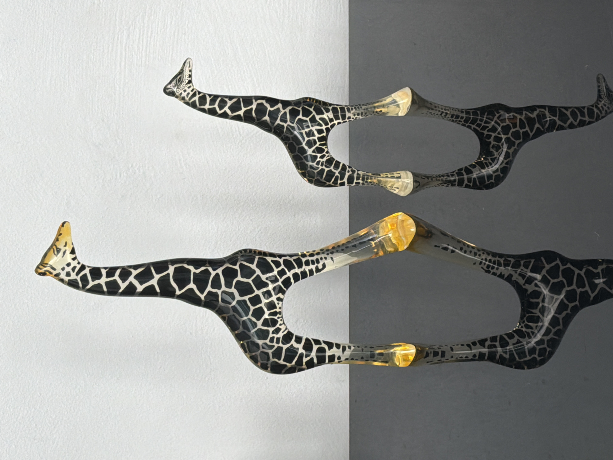 The image displays two abstract, stylized objects against a vertically divided background. The objects exhibit a distinctive giraffe-like pattern of irregular dark shapes on a lighter, glossy surface, suggesting either painted material or a textured finish. Each object consists of an elongated, curved form that terminates in a pointed, slightly upturned end, resembling a giraffe's head and neck. The main patterned body transitions into translucent, amber-toned sections that appear to be made of resin or glass, forming the central and rightmost parts of the structure.
The two objects are positioned horizontally, one above the other, with a noticeable gap between them. The top object appears to be a mirror image or a closely related piece to the bottom one. They extend from the left side of the frame towards the right.
The background on the left side is a light, textured wall or panel, possibly white or light grey, showing a subtle stucco or plaster-like finish. On the right side, the background is a smooth, darker surface, likely a dark grey wall or a shadow effect created by lighting, indicating a sharp edge where the two surfaces meet. The objects cast soft shadows on the lighter background, suggesting ambient or diffused lighting.
There are no people or specific activities depicted. The objects are static, functioning as decorative art pieces, sculptures, or possibly unique architectural elements like handles or hooks, designed with an animalistic motif. No text is visible in the image. The scene conveys an indoor setting, likely a gallery or a well-lit interior space.