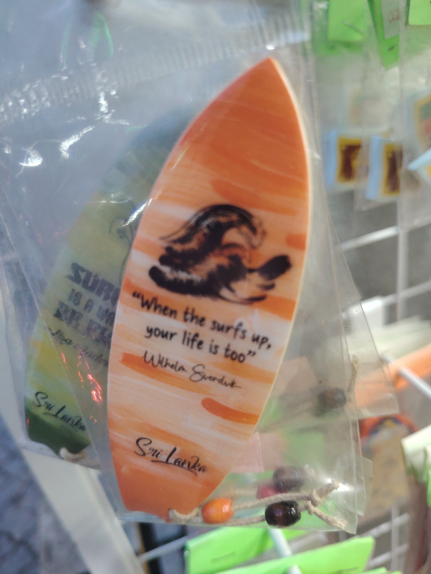 The image shows two miniature surfboards, one orange and one yellow-green, inside clear plastic packaging. Both surfboards have the text "Sri Lanka" written on them in cursive. The orange surfboard has a black and brown abstract design of what appears to be a wave or animal and a quote in black text: "When the surfs up, your life is too" followed by a signature that appears to read "Wilhelm Sverdlik". The yellow-green surfboard has text in black that is partially obscured but reads "SURF IS A WAVE THAT FREE...". Both surfboards have a string with beads attached to the bottom. The surfboards are hanging in what appears to be a market or souvenir shop, with other items visible in the background, including small square objects with yellow and brown markings. The overall lighting suggests an outdoor or well-lit indoor environment.