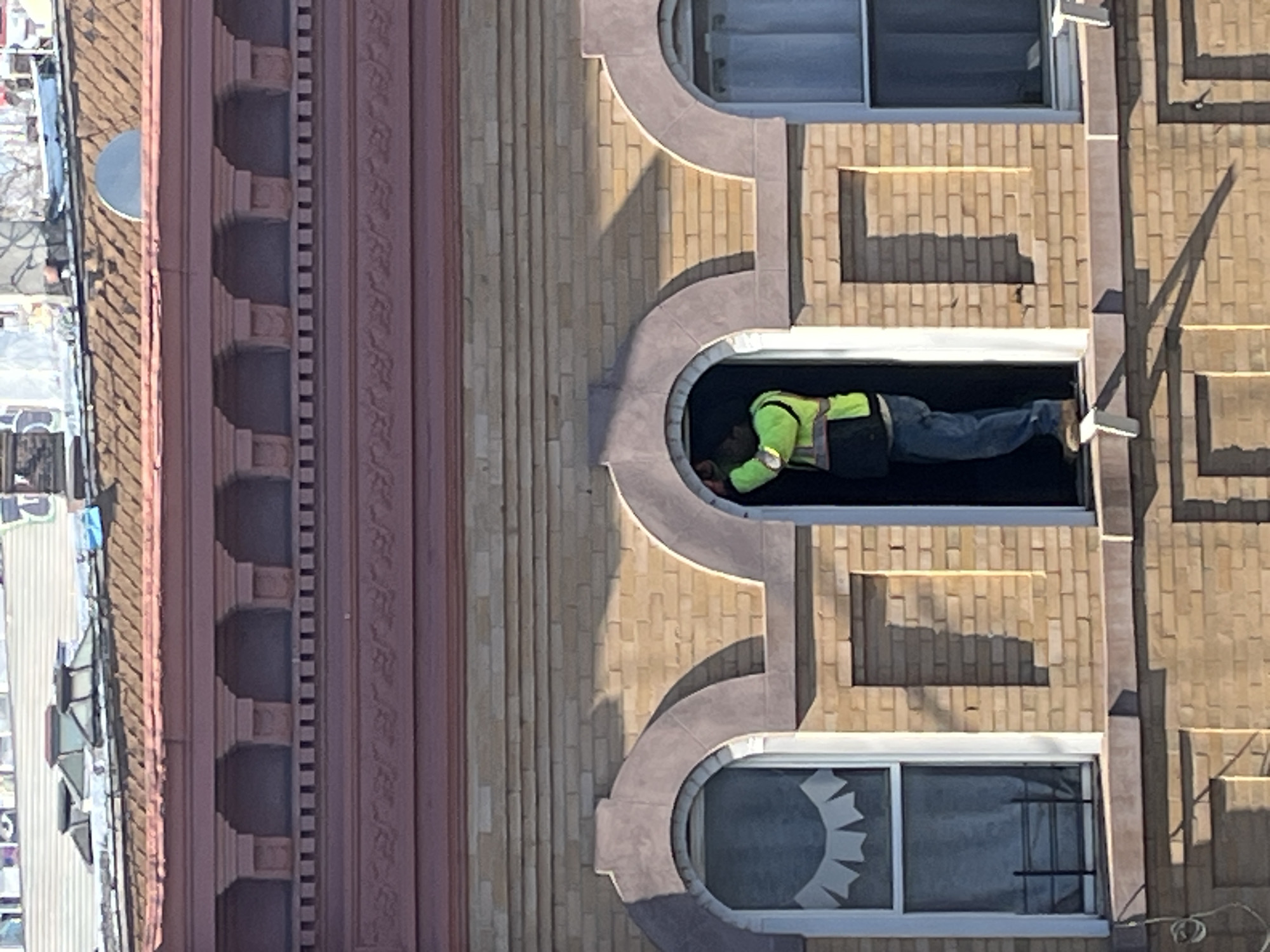 The image displays the upper floors of a light brown brick building, characteristic of urban architecture in New York, United States of America. The exact location within New York City is not discernible.
A male individual is positioned within an arched window opening, standing on the sill. He wears a neon yellow-green high-visibility vest over dark clothing, blue jeans, and tan work boots. His arms are raised, and his hands are engaged with the upper section of the white window frame, suggesting active work or manipulation.
To the left of the individual, a closed arched window, also with a white frame, displays sheer white curtains and a decorative white paper garland on the upper pane. A black metal security grate is visible externally on its lower section. A similar arched window, partially visible to the right, also contains sheer white curtains.
The building facade above the windows features a reddish-brown decorative cornice with multiple arched niches and a patterned frieze. Above this, a sloped, brown-shingled roof is visible, along with a grey satellite dish and other rooftop structures. Direct sunlight illuminates the scene, casting shadows across the brickwork.