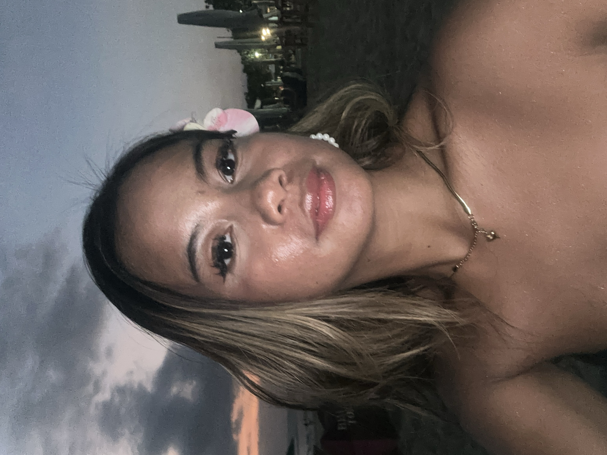 A medium shot shows a woman with shoulder-length highlighted brown hair and light brown skin. She has a pink flower in her hair, along with a pearl earring and a delicate gold necklace. The background appears to be an outdoor scene in Kuta, Indonesia, as evidenced by the architecture and tropical vegetation in the distance. It is dusk or night, as indicated by the dim lighting and visible cloud cover in the sky.