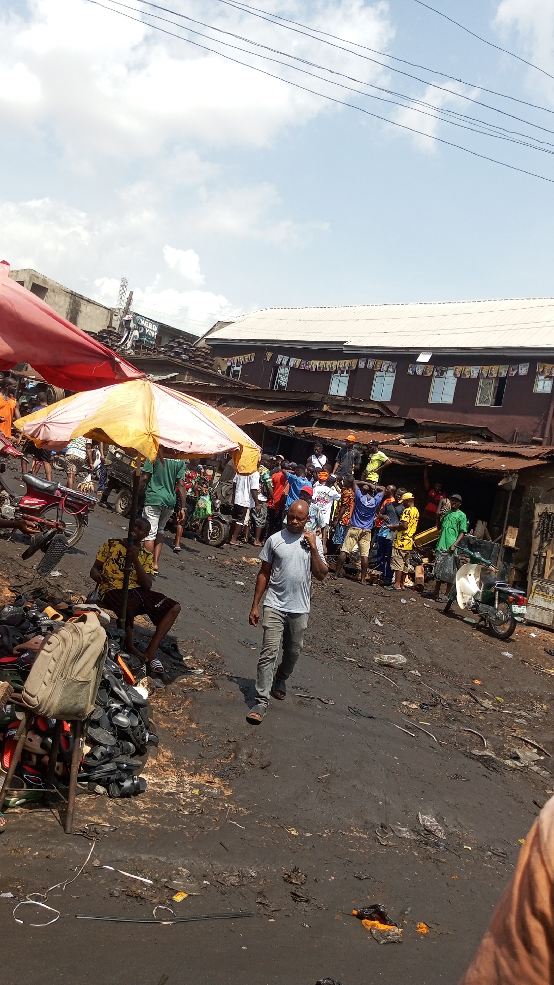 This image captures a vibrant, bustling outdoor market scene in Onitsha, Nigeria, under bright daylight with a partly cloudy sky. The ground is dark, appearing muddy or heavily soiled, with scattered debris.
In the foreground, a man in a grey t-shirt and dark trousers walks towards the camera. To his left, another man sits under a yellow and white market umbrella, surrounded by large piles of sandals and shoes, with a backpack resting nearby. Further back, a red market canopy is partially visible on the left. Several motorcycles or mopeds are parked or moving through the scene.
A large crowd of people is gathered in the mid-ground, many standing or interacting in front of market stalls with corrugated iron roofs. The atmosphere suggests a typical day of commerce and activity. In the background, multi-story buildings line the market, with banners or decorative elements strung across the upper level of one structure. Partially visible text on one banner reads "NEED PATH". The overall setting conveys a dynamic urban market environment.