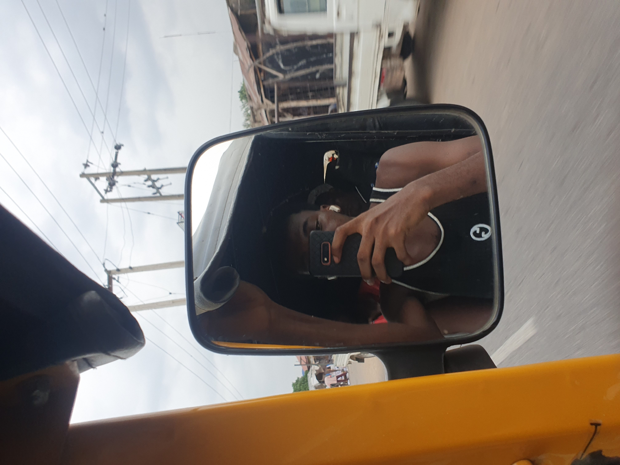 An upward-angled, candid photograph captures a reflection of a person taking a selfie in a vehicle's side-view mirror, set against the backdrop of a street scene in what appears to be Lokoja, Nigeria.
The central subject is a dark-skinned individual, likely male, reflected in the mirror. They are wearing a black tank top with white striped accents and appear to have white earbuds in their ears. With their right hand, they hold a black smartphone, featuring a reddish outline around its camera module, positioned to take a picture of their reflection. Their face is partially obscured by the phone.
The mirror itself is rectangular with a black frame, attached to a bright yellow vehicle body, suggestive of a "Keke Napep" (motorized rickshaw) or a similar type of commercial transport common in the region. A small white logo or symbol, resembling a stylized 'e' or 'C' within a circle, is visible on the lower part of the mirror's frame.
Outside the vehicle, the background shows a dynamic street view, blurred due to motion, indicating the vehicle is moving. In the upper left quadrant, utility poles with numerous overhead wires cut across an overcast, cloudy sky. Further into the background, other vehicles, possibly a white truck or bus, and several simple buildings or structures are visible. The overall setting depicts a busy, perhaps urban or semi-urban, environment during daytime with diffused light, suggesting an overcast sky.