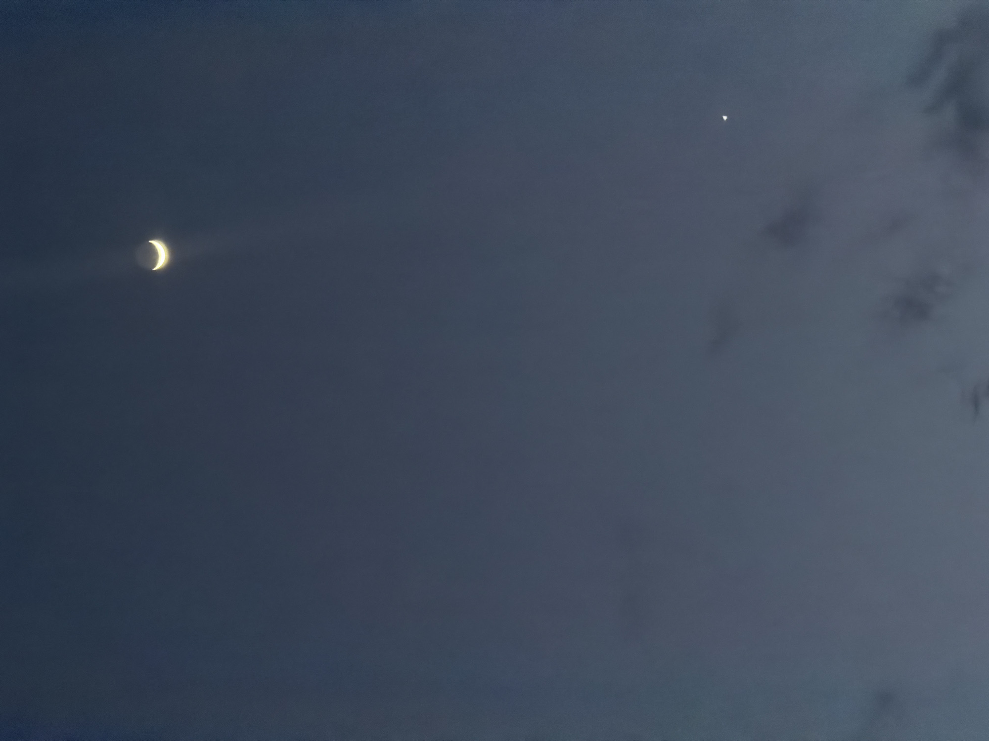 The image shows a dark blue sky at dusk or night. A crescent moon is visible in the upper right quadrant of the image, emitting a faint glow. Below and to the right of the moon, a small, bright object, possibly a planet or star, is visible. There are faint cloud formations scattered throughout the sky. The image was taken in Bukuru, Nigeria.