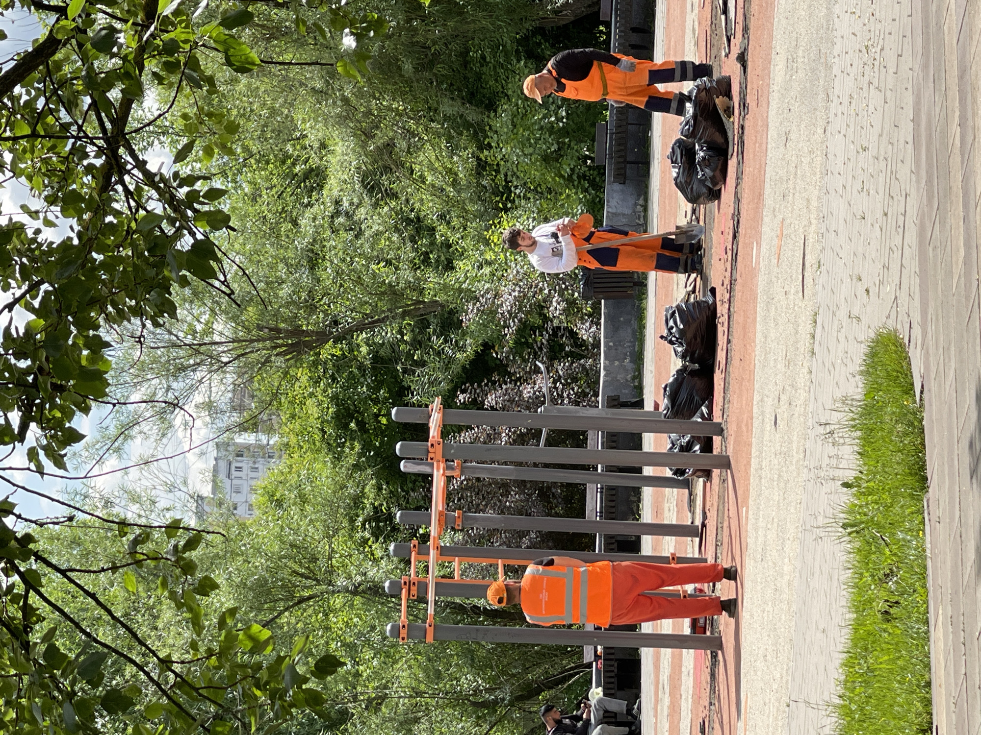 The image depicts a scene in a park in Moscow, Russia. Three workers wearing orange uniforms are cleaning the park. They are picking up trash and placing it in black plastic bags. There are bags of trash on the pavement next to a wrought-iron fence and fitness equipment. The park has lush greenery, with trees and grass visible in the background. A building can be seen through the trees in the distance.