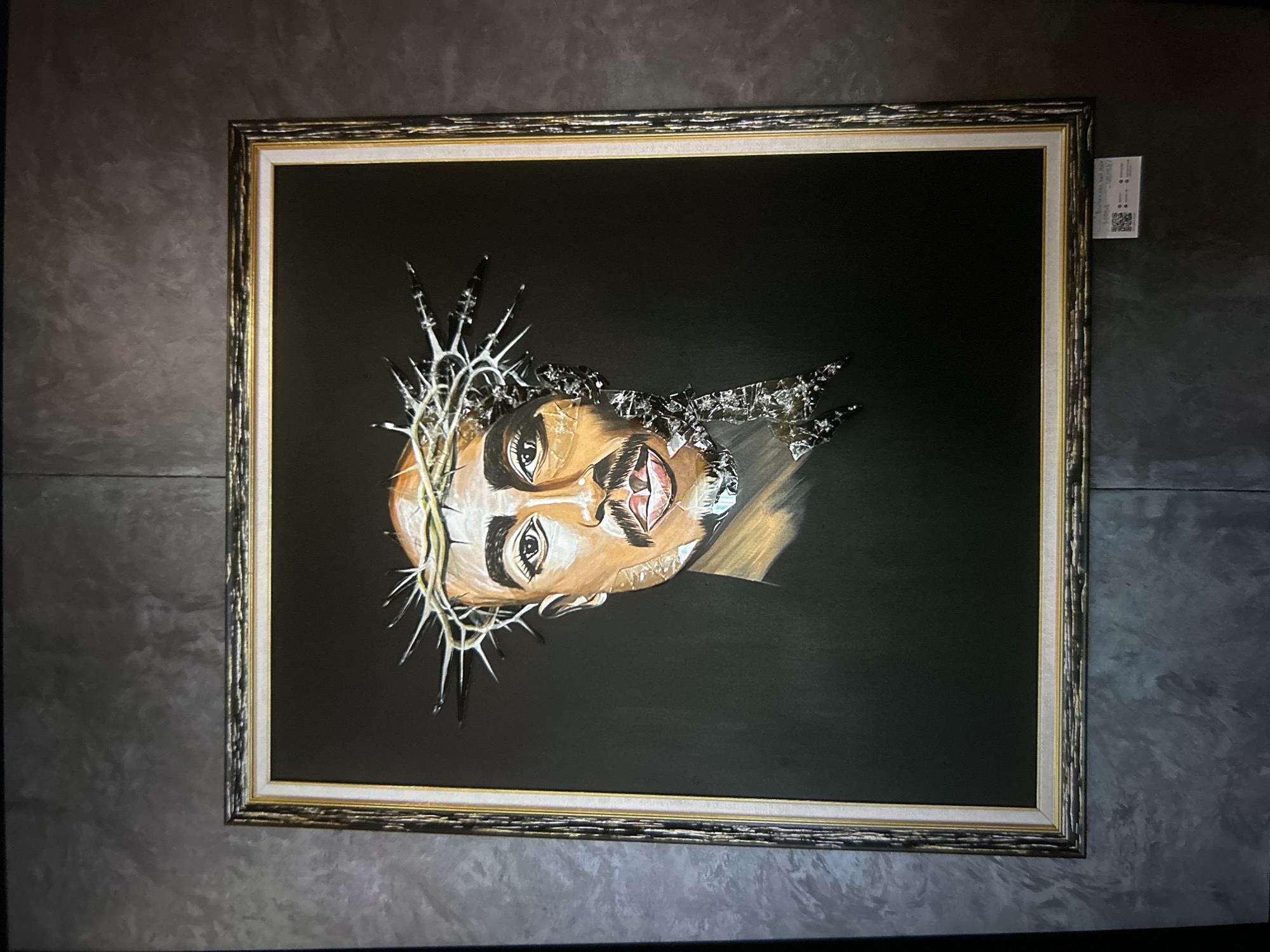 An framed artwork hangs centrally on a dark grey, textured wall in the city of Unknown, Unknown. The artwork depicts a portrait of a man, identifiable as Tupac Shakur, wearing a crown of thorns. The left side of the man's face (viewer's right) is rendered in painted tones, while the right side (viewer's left) appears to shatter into numerous metallic, reflective fragments, extending from the crown of thorns downwards. The background of the artwork is solid black. The piece is enclosed within a multi-layered frame consisting of an inner white matboard, followed by a gold-colored border, and an outer dark, ornate frame with gold accents. Below the bottom right corner of the frame, a small white informational plaque featuring two QR codes and obscured text is affixed to the wall. The wall itself exhibits a mottled, uneven dark grey texture, possibly indicative of concrete or stucco.