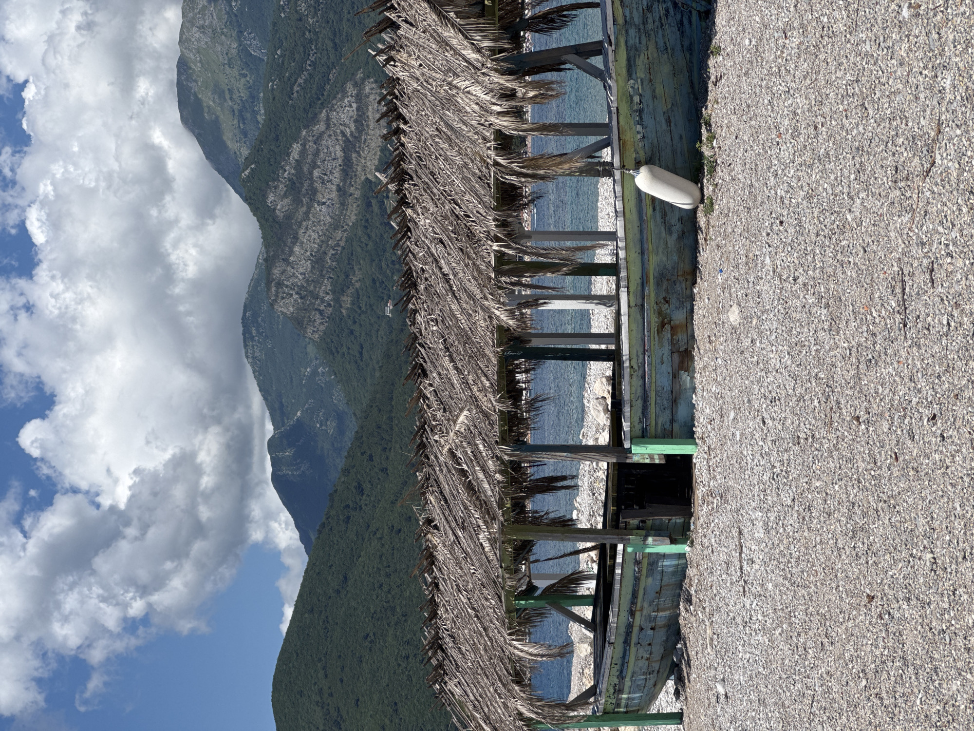 This image captures a serene coastal landscape in Budva, Montenegro, under a bright daytime sky. Dominating the background are majestic mountains, displaying a rugged texture with areas of dense green vegetation contrasting with rocky grey surfaces, rising towards a clear blue sky interspersed with fluffy white cumulus clouds.
In the midground, the calm, deep blue sea stretches out, meeting a pebble beach. A rustic wooden structure, likely a beach bar or shelter, extends across the middle of the frame. Its roof is made of dried palm fronds or similar thatched material, giving it a natural, weathered appearance. The wooden framework of the structure is painted in faded hues of green and blue, suggesting parts of an old boat hull might have been repurposed. A white boat fender or buoy is visible, tethered to the structure and resting on the pebbles.
The foreground is filled with a wide expanse of light grey and beige pebble beach, comprised of small stones and shell fragments, indicating a natural coastal environment. The bright sunlight illuminates the scene, creating distinct shadows and highlighting the varied textures of the mountains, the thatched roof, and the pebbles. The absence of people lends a tranquil and undisturbed atmosphere to the scene. There is no visible text in the image.