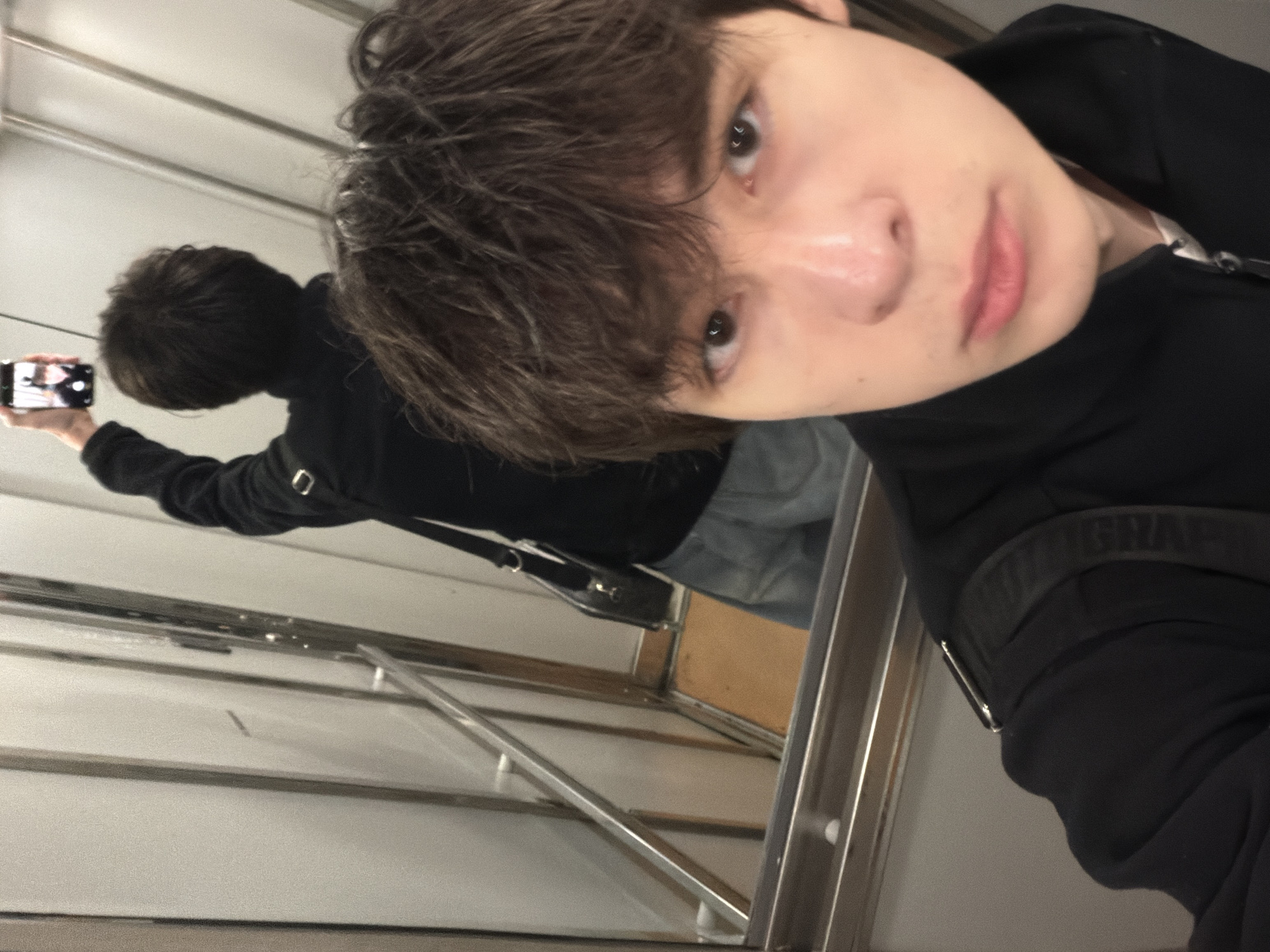 A young person with dark, messy hair takes a selfie in an elevator. The person is wearing a black jacket and light blue jeans. In the background, their reflection is visible, holding a smartphone to take the picture. The elevator has metal walls and a light-colored floor. The overall impression is of a casual moment captured in an urban setting.