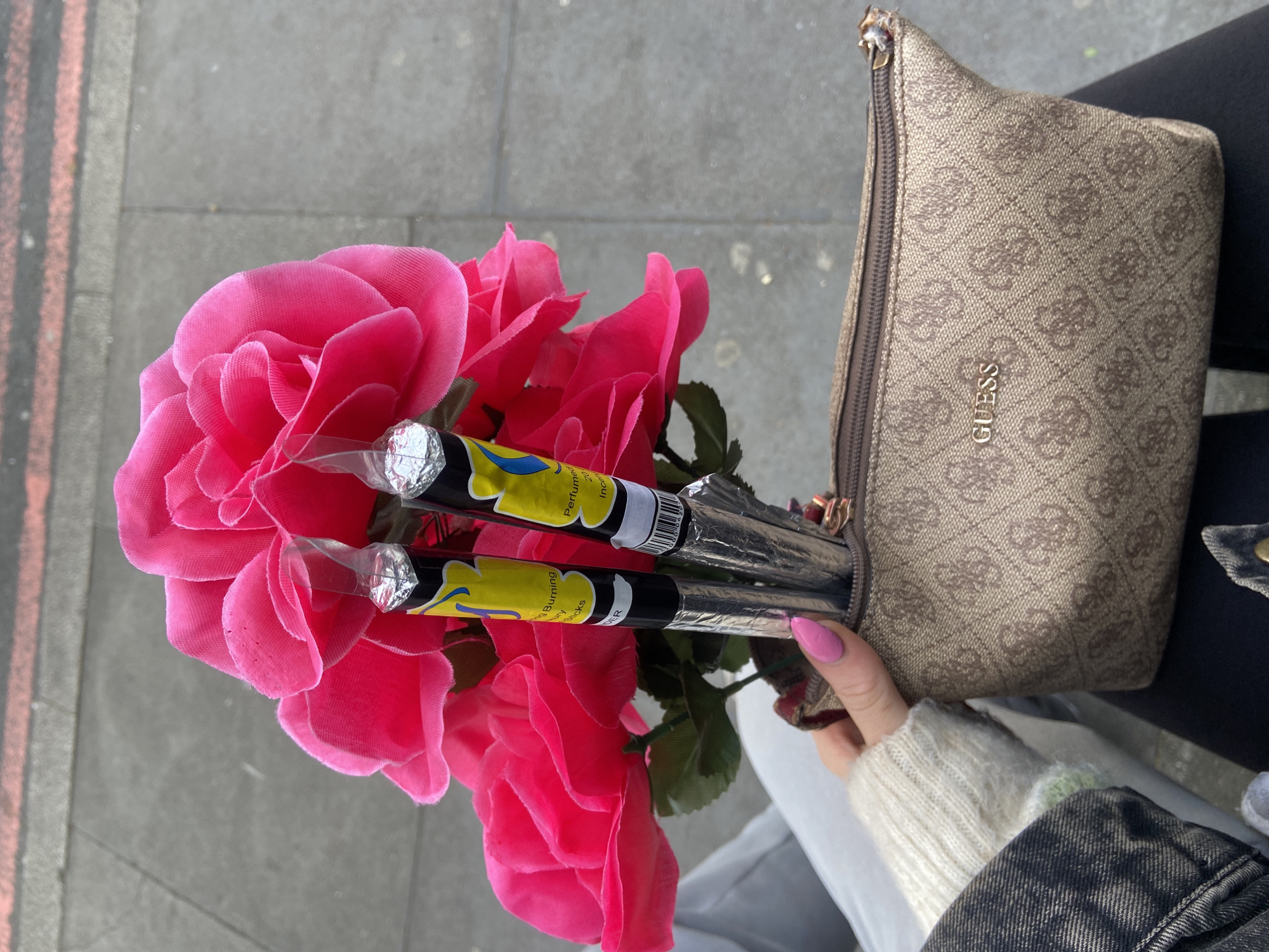 Here is a forensic description of the image:
The image shows a person holding a bouquet of large, artificial pink roses along with two wrapped incense sticks. The incense sticks are silver wrapped with a yellow label that reads "Burning Luxury Sticks". The person is also holding a tan-colored "Guess" brand purse, partially open, suggesting that the roses and incense sticks are being placed inside. The person's hand, with pink painted nails, is visible. They are wearing a cream-colored sweater with a hint of green and a denim jacket. The background shows a sidewalk or street with a hint of black/orange lane markings. The location cannot be determined to be in the City of London based on visual evidence alone.
