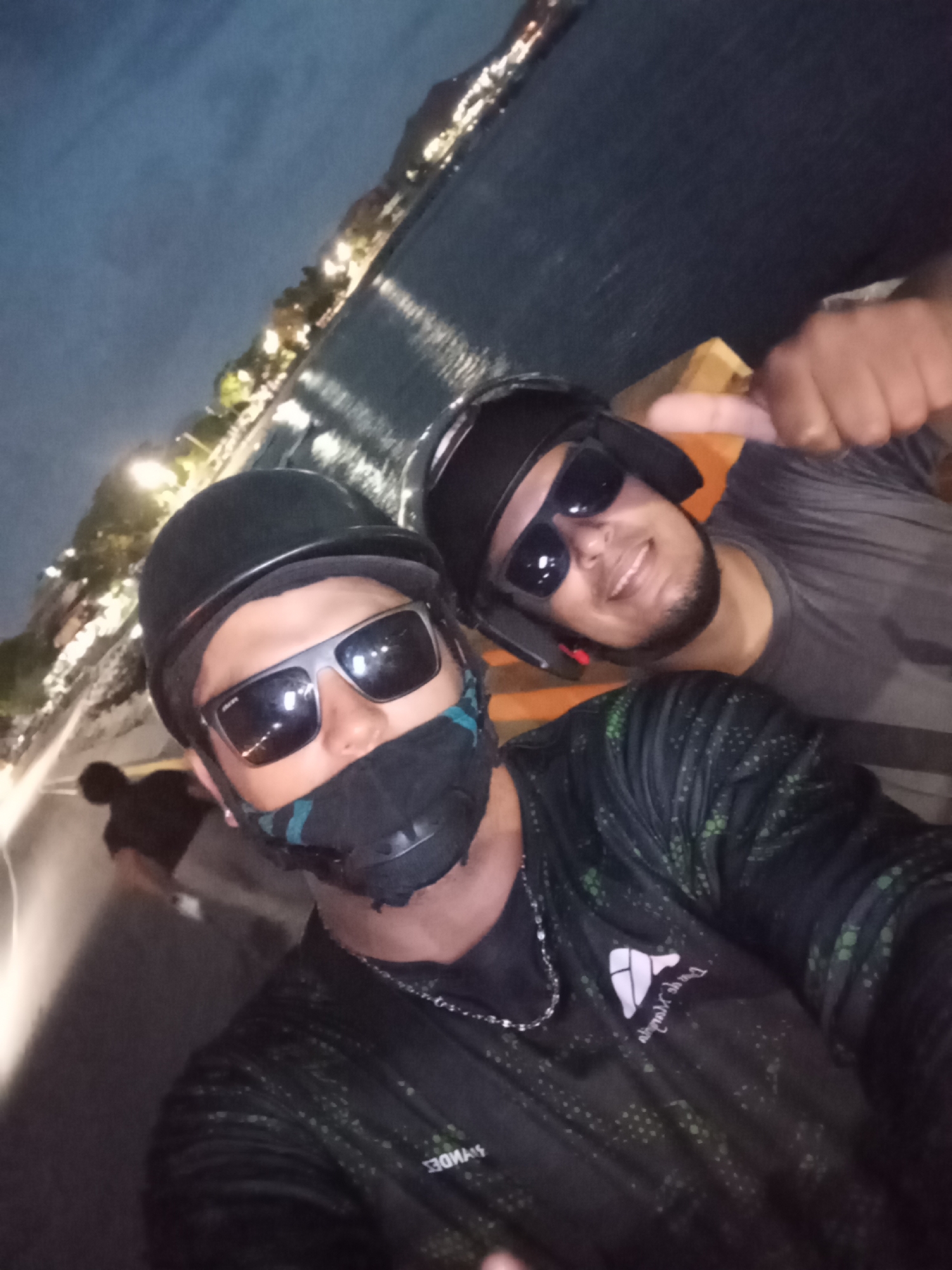 The image is a selfie taken at night, likely on a street or boardwalk with a body of water and a lit-up coastline in the background. Two individuals are visible, both wearing helmets and face coverings. The person in the foreground is wearing sunglasses and a dark, patterned shirt. They are looking at the camera and appear to be making a gesture with their hand. The person in the background is also wearing a helmet and sunglasses, and is giving a thumbs-up gesture. The overall atmosphere suggests a leisurely outing. The presence of a lit-up coastline with buildings is consistent with an urban or semi-urban setting at night. Given the context of Juan Griego, Venezuela, this scene could depict a nighttime stroll or ride along the waterfront in the town.