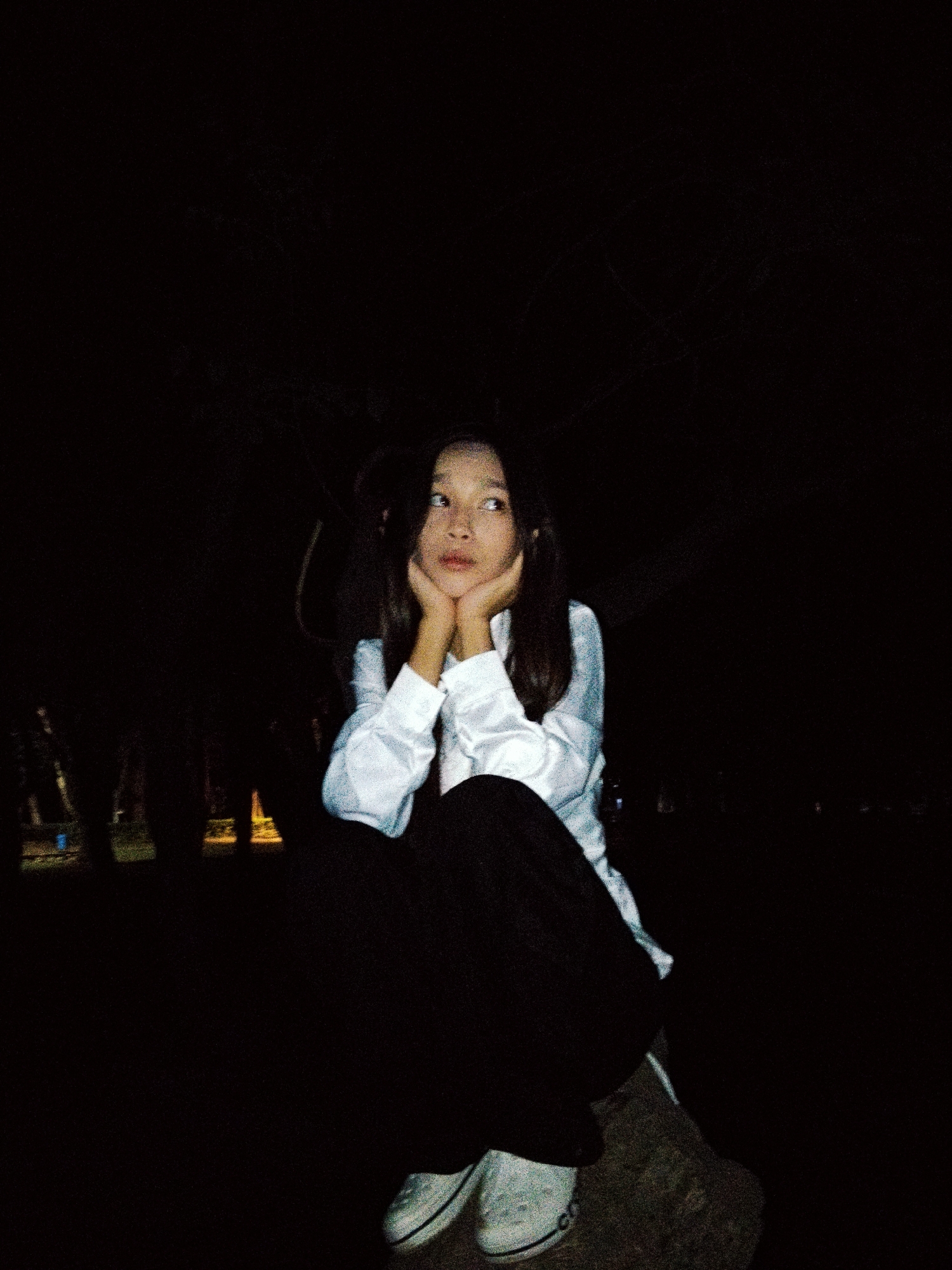 A young Asian female is depicted in a low-angle shot, crouched down in what appears to be an outdoor setting at night. She is wearing a white collared shirt and dark pants, with white sneakers. Her hands are clasped beneath her chin, and she is looking to her right, with a thoughtful or pensive expression. The background is dark, with some indistinct lights and silhouettes of trees visible, suggesting a park or wooded area. The overall lighting is dim, with the subject illuminated by a light source from the front, casting subtle shadows. The location is identified as Bishkek City, Kyrgyzstan.