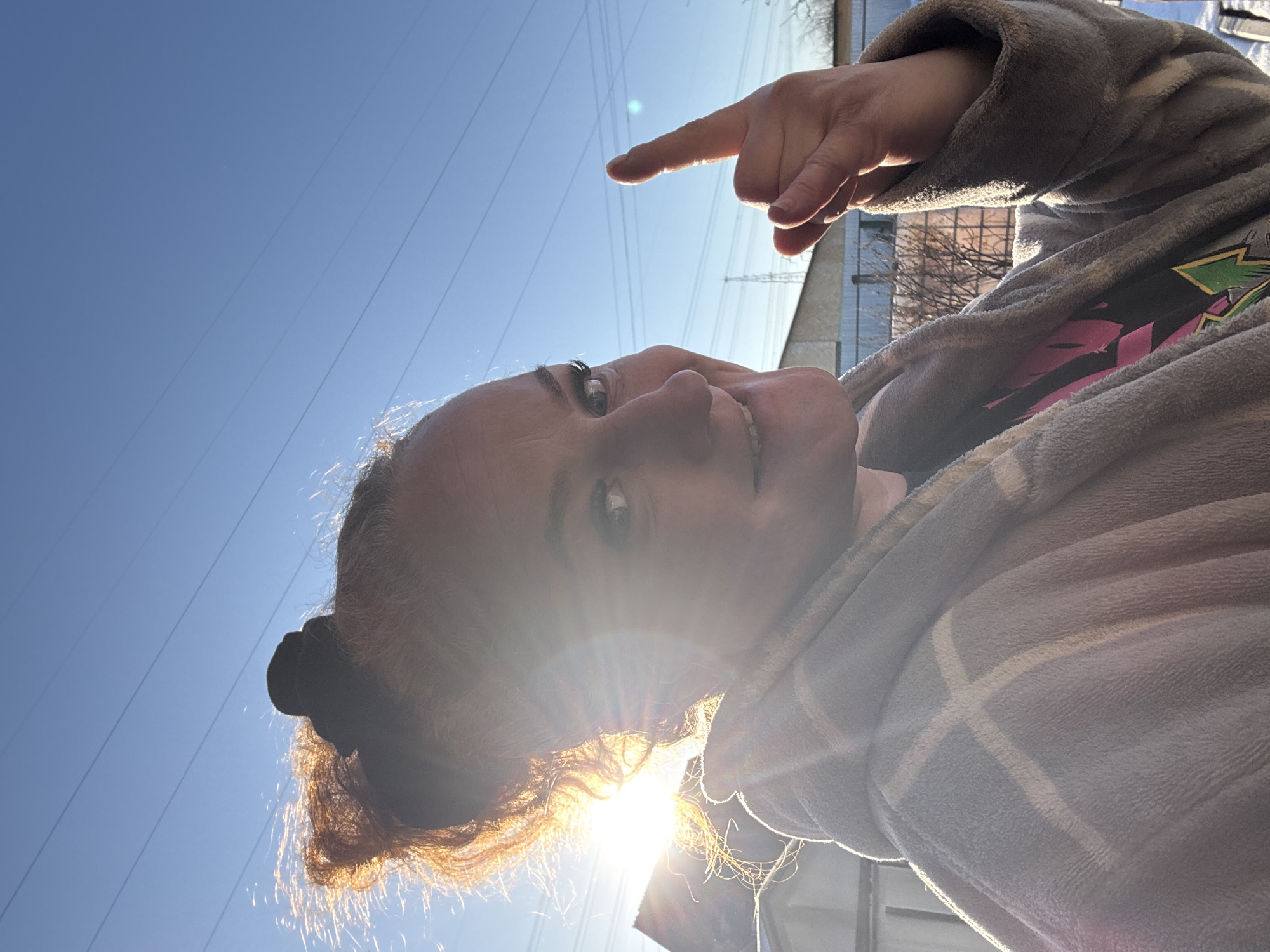 A person is facing the camera and pointing upwards with their index finger. The background consists of a clear blue sky with power lines. The sun is low, causing lens flare. A building is visible behind the person, and a tree with bare branches is next to it. The person is wearing a grey robe. The picture may have been taken outside, based on the lighting and background elements. The location is the city of Chaikovsky, Russia.