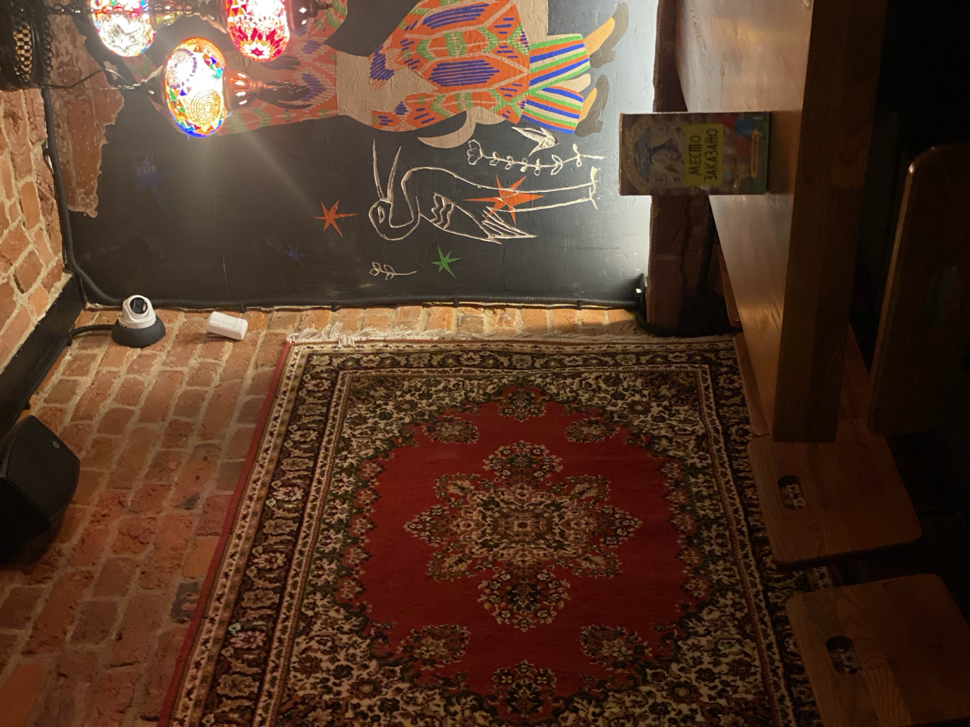 This image captures the interior of an establishment, identified as being in Saint Petersburg, Russia by the Russian text visible. The scene displays an eclectic decor featuring an exposed red brick wall on the left, adorned with a large, intricately patterned red carpet hanging vertically. High on the brick wall, a black speaker is mounted. Near the upper left corner, a white dome-style security camera and a smaller white rectangular sensor are affixed to the brickwork, with black conduits running alongside.
The adjacent wall on the right is painted a dark color (likely black) and features a colorful mural. The mural depicts the lower half of a stylized human figure wearing a vibrant, patterned skirt or dress in orange, blue, green, and white, along with black shoes. A white outline drawing of a stork or crane-like bird is also part of the mural, accompanied by various star shapes and plant motifs in green, orange, and white. Above this mural, multiple ornate, mosaic-style glass lamps with metallic accents hang, casting colorful light.
In the foreground, a polished wooden table extends from the center to the right. Several simple wooden chairs or stools are partially visible around the table. On the table, a small, upright sign displays the Russian text "МЕСТО ЗАКАЗАНО" (Mesto Zakazano), meaning "Place Reserved," on a yellow background, accompanied by colorful illustrations. No individuals are present in the image.