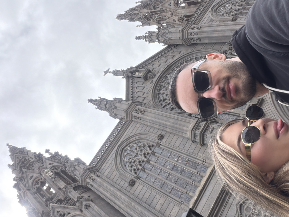 Two individuals are depicted in a selfie taken in front of the Church of San Juan Bautista in Arucas, Gran Canaria, Spain. The male subject, positioned on the right, has short dark hair and a beard, wears square grey-framed sunglasses, and a dark hooded sweatshirt. He looks directly at the camera. The female subject, positioned on the left, has long blonde hair with highlights, wears round gold-framed sunglasses, and is shown with her tongue extended playfully. Both individuals exhibit light skin tones and appear to be in their 20s or 30s. The background is dominated by the monumental Neo-Gothic architecture of the church, constructed from dark volcanic stone, featuring a tall, ornate spire, pointed arches, intricate tracery, and stained-glass windows. The sky overhead is overcast and grey.