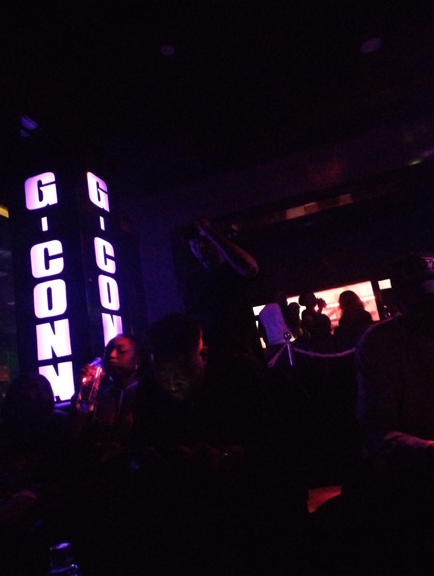 The image depicts the interior of what appears to be a nightclub or lounge, indicated by the brightly lit "G-CONY" sign in purple hues on the left. People are visible, some seated at tables, others standing near a rope barrier or line. A woman is seen holding a glass, possibly a drink. The overall ambiance is dark, typical of such establishments, with dim lighting. The location is presumed to be in Gombe, Nigeria, based on the place mentioned in the request.