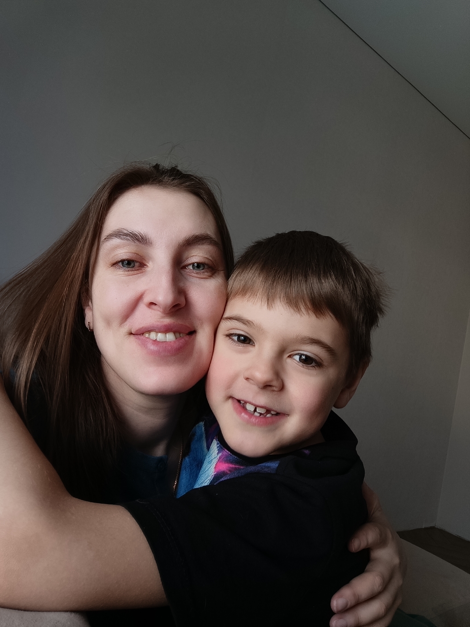 A selfie taken indoors features a woman and a young boy. The woman has long, brown hair and blue eyes, and is smiling. She is wearing a blue shirt and small hoop earrings. The boy has brown hair and brown eyes, and is also smiling. He is wearing a black hoodie with a colorful design underneath. The woman is hugging the boy. The background is a plain, gray wall with a corner visible on the right side. The location is the city of Chaikovsky, Russia.