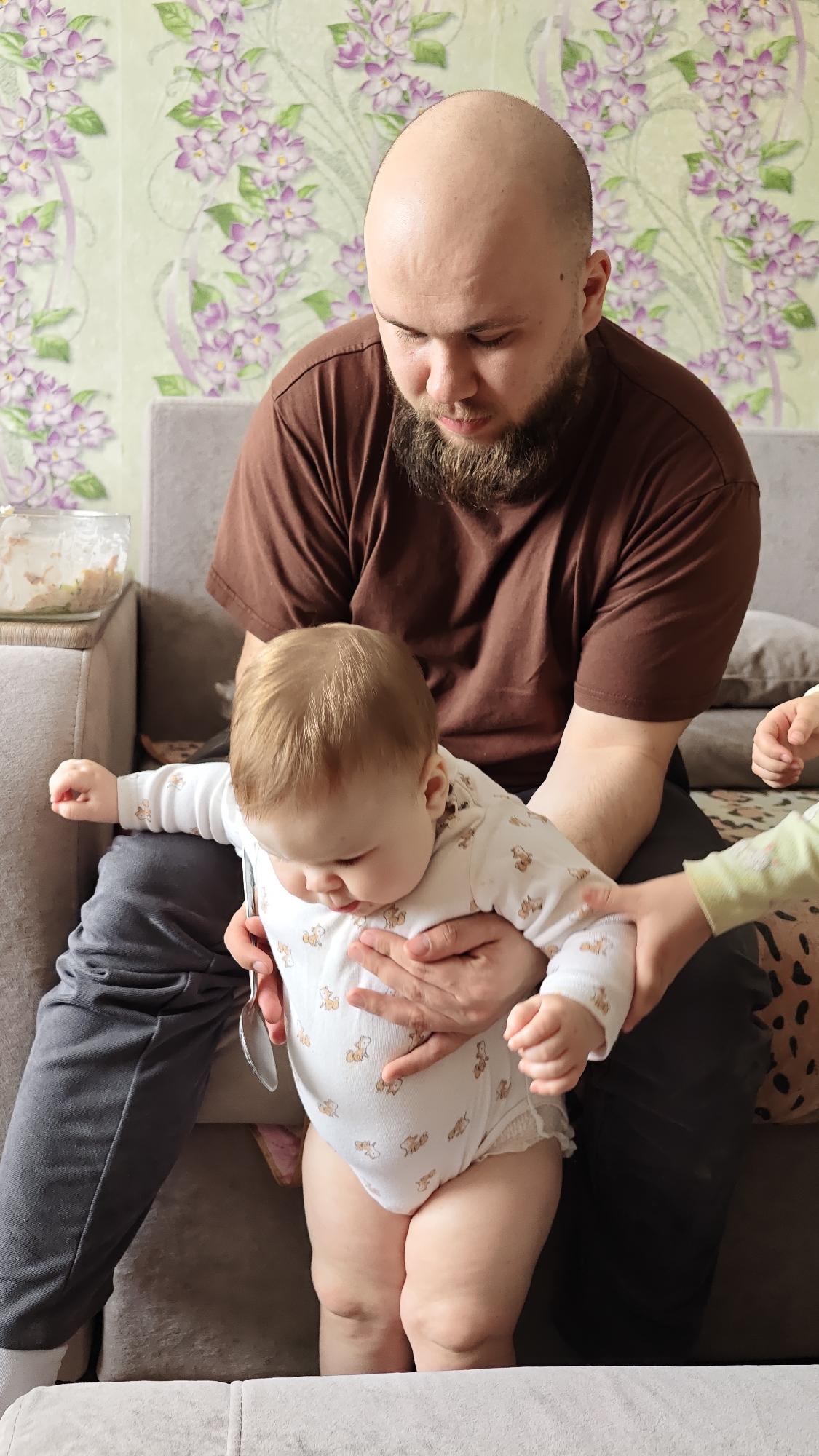 The image depicts an indoor scene featuring a bald man with a beard sitting on a grey couch. He is wearing a brown T-shirt and grey pants. He holds a baby in his lap who is wearing a white onesie with a pattern of small animals. The baby is standing and the man is supporting the baby. To the right of the baby is the arm of a child wearing a light green shirt. Behind the couch is a wall with a floral pattern. To the left of the man is a glass bowl containing food or snacks. The scene may be located in Уральское, Russia.