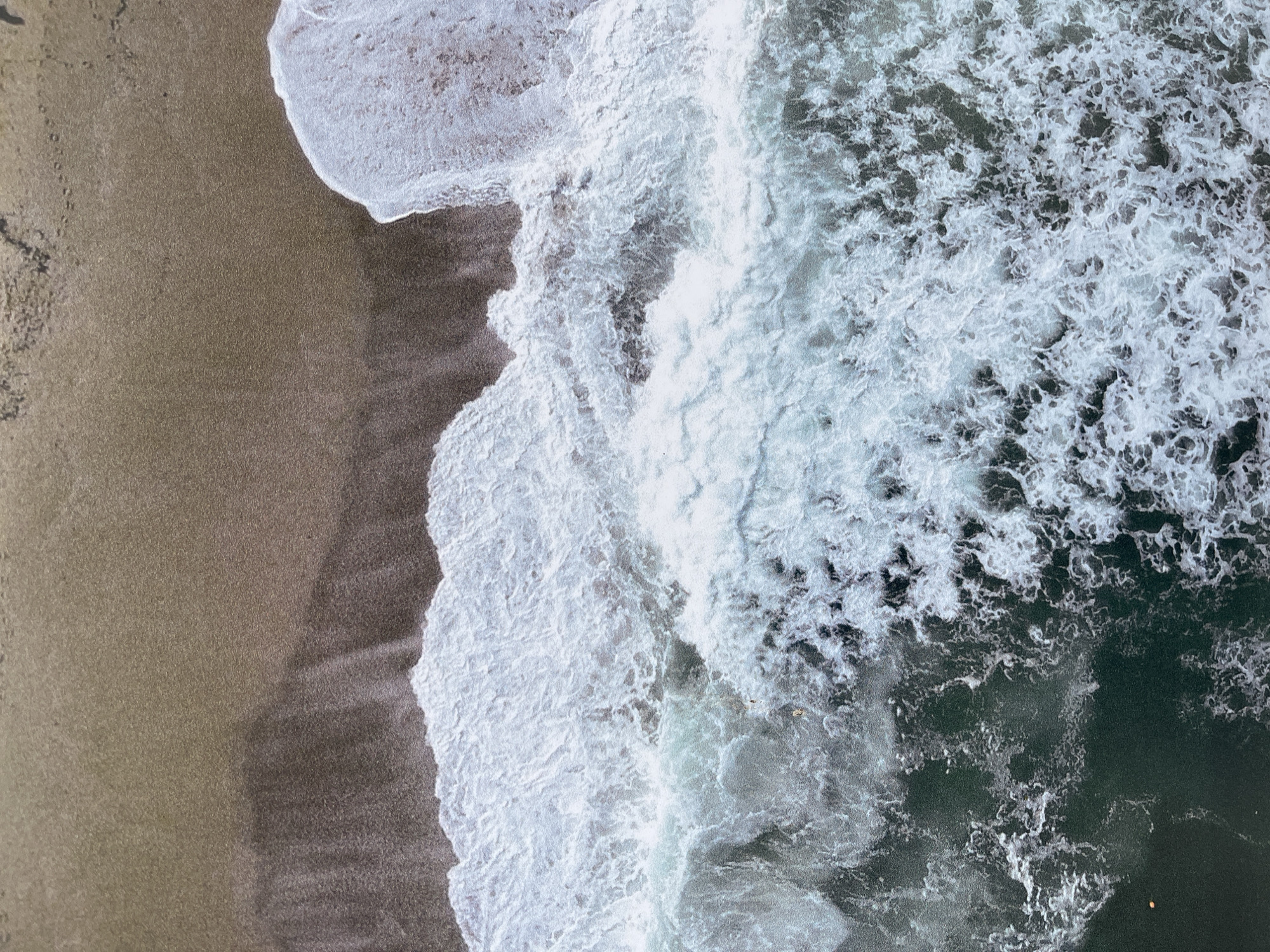 This is an aerial photograph of a beach and ocean shoreline, featuring sand and turbulent water. The sand is on the left side of the image, showing a brownish-grey color with subtle variations in tone, with some spots that indicate recent contact with water. Moving to the right, the water transitions from dark green-blue to white as it churns near the shore, forming foamy wave patterns. The frothy texture of the white waves contrasts sharply with the smoother texture of the sand and deeper water. The scene likely depicts a shoreline with active wave action, suggesting a dynamic coastal environment. While the image does not provide specific location information, the provided context specifies Burgas, Bulgaria. Based on the coastal features, the scene is presumed to be located on a beach along the Black Sea in the Burgas region, however the image does not show any elements that would indicate a specific location within the city.