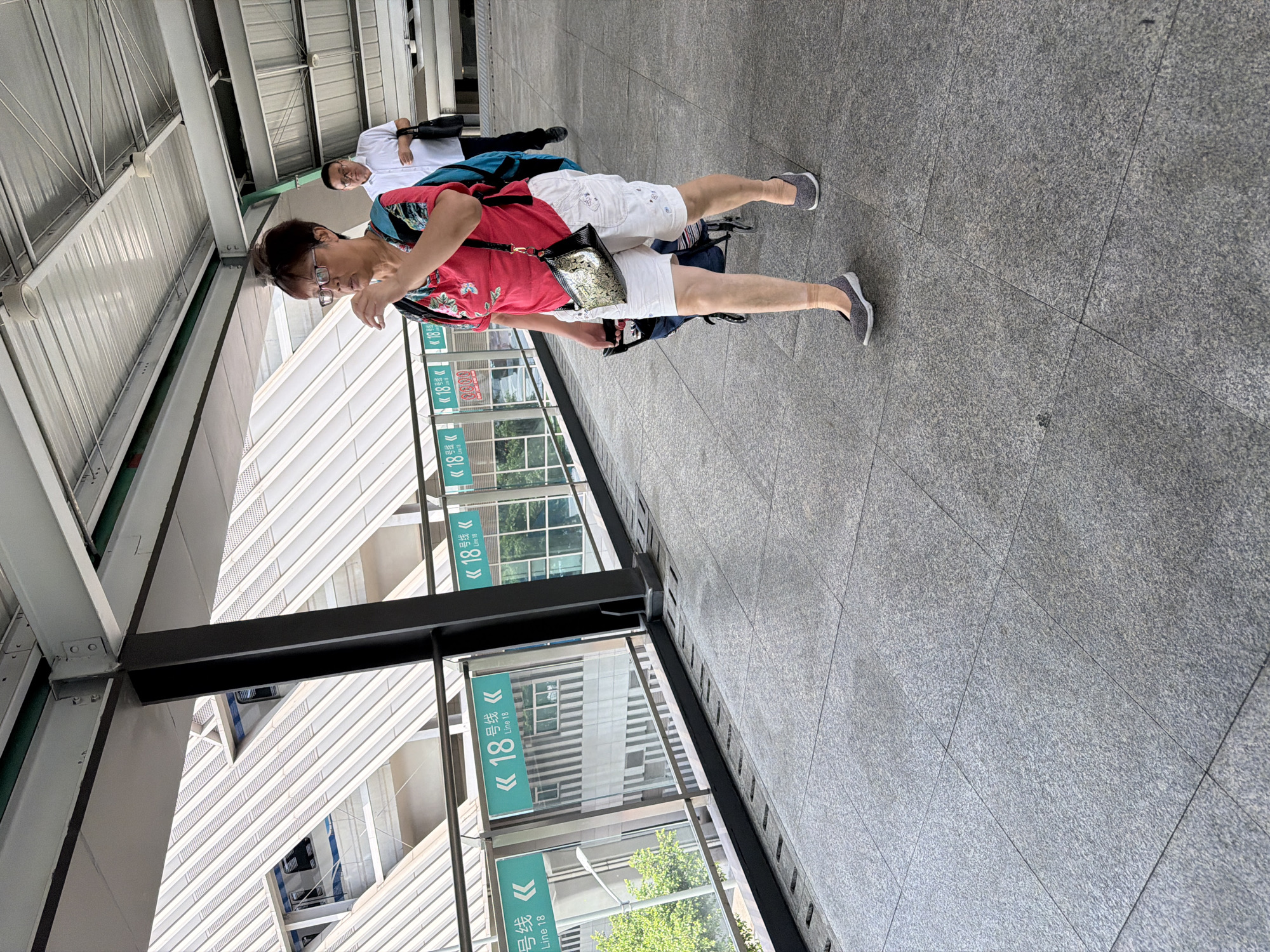 An overhead shot captures a woman in a bright pink floral t-shirt and white shorts walking on a paved walkway. She is wearing glasses and has her hair pulled back. A patterned crossbody bag is slung over her shoulder, and she is pulling a small rolling cart. In the background, a man in a white shirt walks away from the camera. The walkway appears to be elevated, with a railing overlooking a street or building. Green signs indicating "Line 18" are visible along the railing. The architecture suggests a modern urban setting, possibly a transit station or a covered pedestrian bridge.