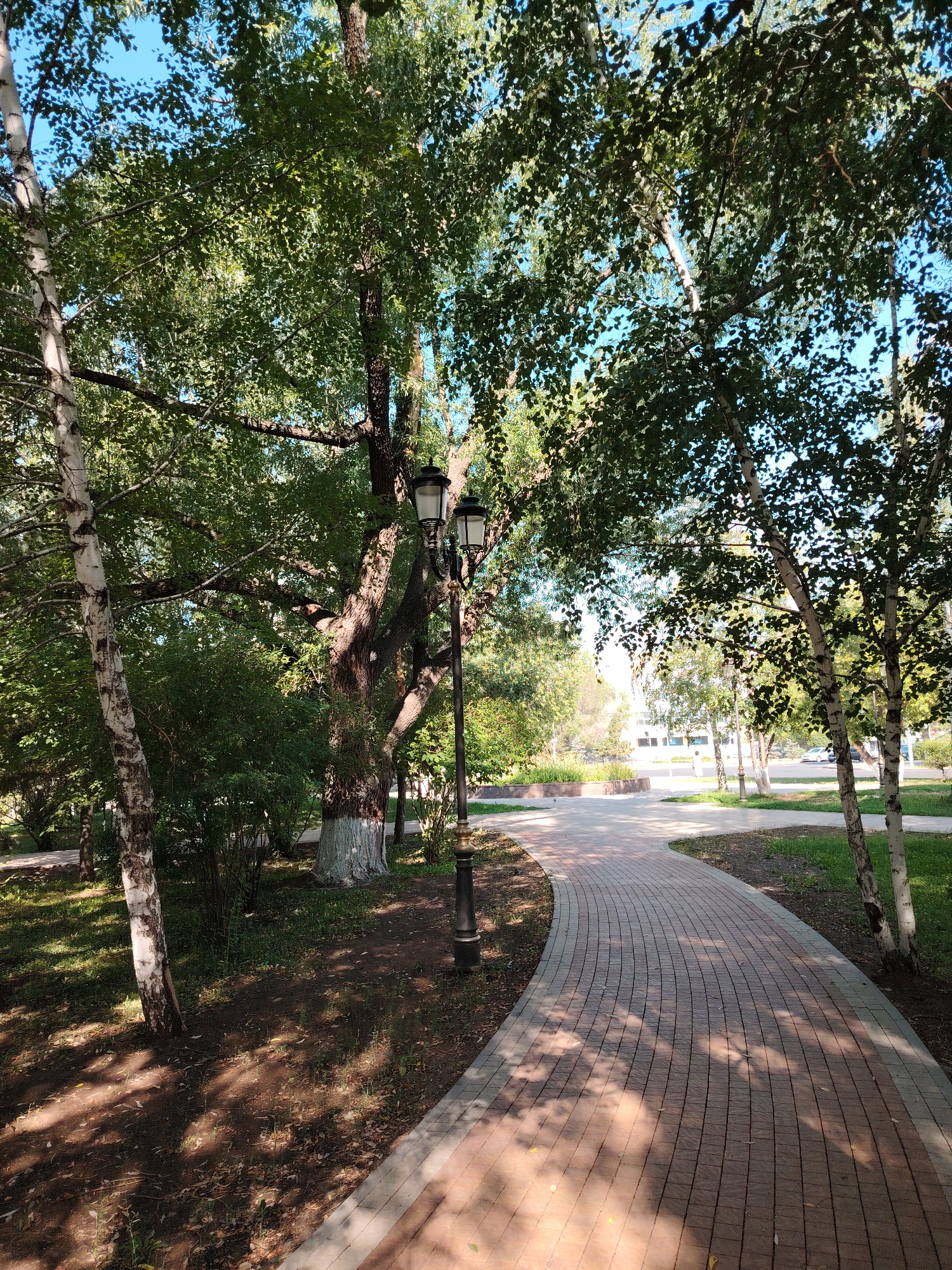 The image depicts a paved pathway winding through a park in Astana, Kazakhstan. The pathway is constructed of red and gray bricks, laid in a curved pattern. Large trees with lush green foliage line the path, casting dappled shadows on the ground. A black lamppost with two ornate lamps stands near the path. In the background, there is a glimpse of buildings and a street with cars, indicating an urban setting. The sky is a clear blue. The scene appears serene and well-maintained, suggesting a public park or a green space within the city. There are no people visible in the image.