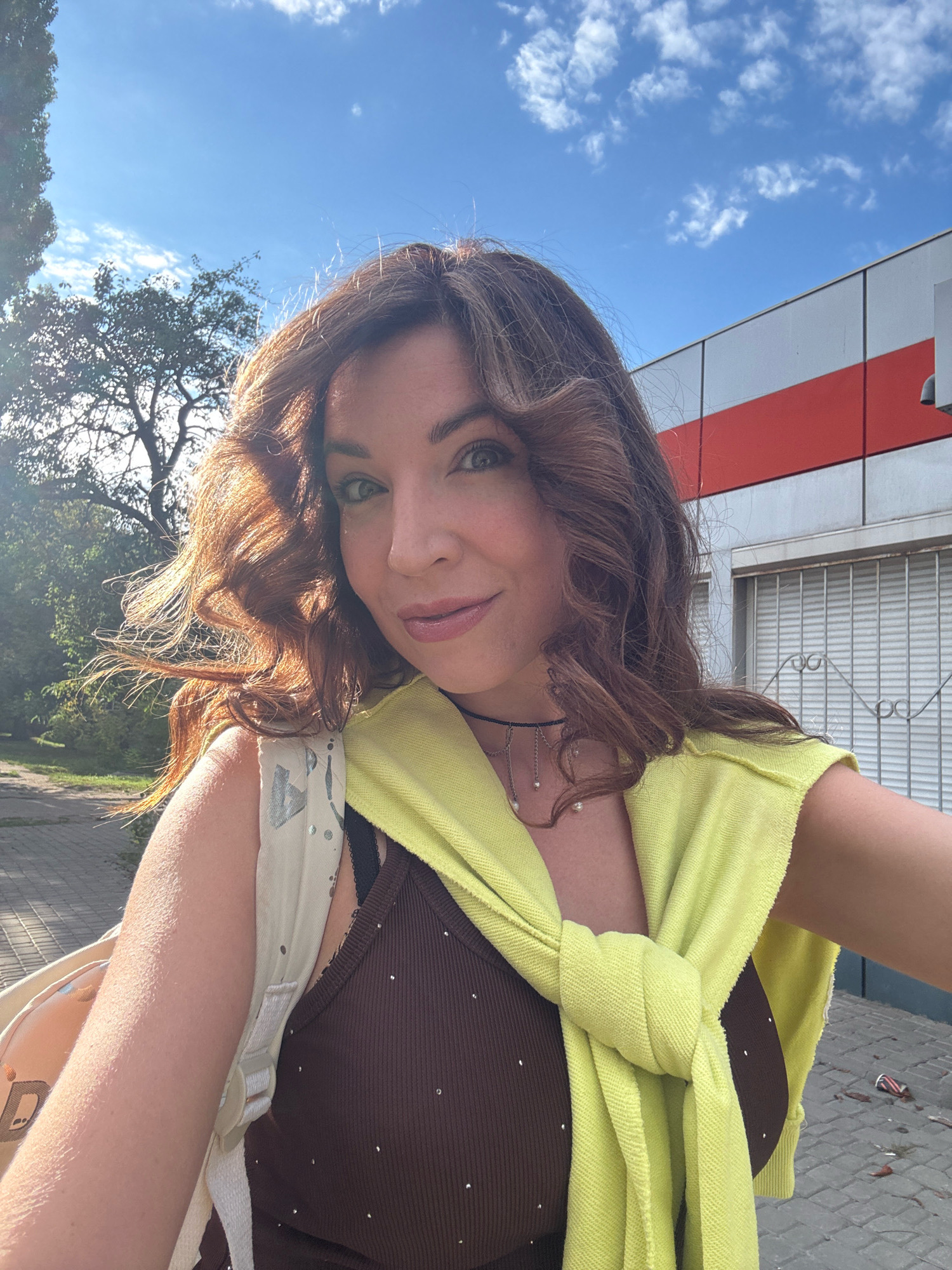 A selfie is taken outdoors on a sunny day in the city of Kryvyi Rih, Ukraine. The subject, a woman with long, wavy brown hair, is positioned in the foreground. She is wearing a brown, sleeveless top adorned with small, reflective embellishments. A bright yellow sweater is tied around her shoulders, with one sleeve draped over her chest. She has on a dark choker necklace with delicate silver chains and pearls. Her left arm is visible, and she is carrying a light-colored backpack with a pattern of abstract shapes and dots.
Behind her, to the left, is a tree with green foliage, and a paved walkway. To the right of the woman is a modern building with horizontal red and white bands. A portion of a light-colored, slatted garage door is visible on the building. The sky above is blue with scattered white clouds. Debris, including what appears to be a crushed can, is scattered on the ground to the right of the building. The lighting suggests it is daytime, possibly late afternoon, given the angle of the sun.