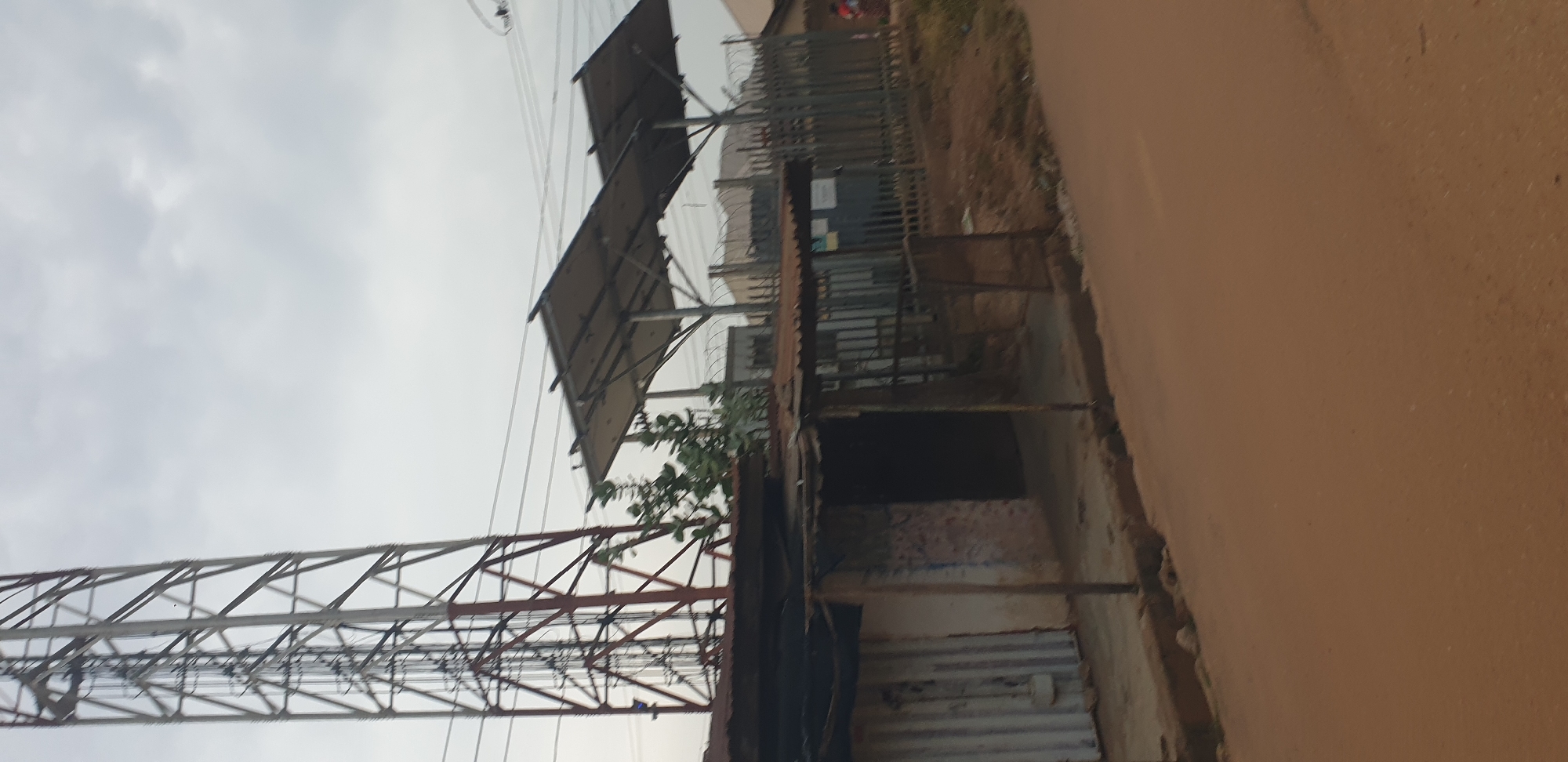 In Sabon Tasha, Nigeria, a cell tower dominates the left side of the frame. Adjacent to it, a rustic, open-fronted stall with a corrugated metal roof and vertical wooden supports stands on a dirt path. Behind the stall, a metal fence with barbed wire stretches across the midground, separating the area from a building with solar panels mounted on an elevated structure. The ground in the foreground is a reddish-brown dirt road. The sky is overcast and cloudy.