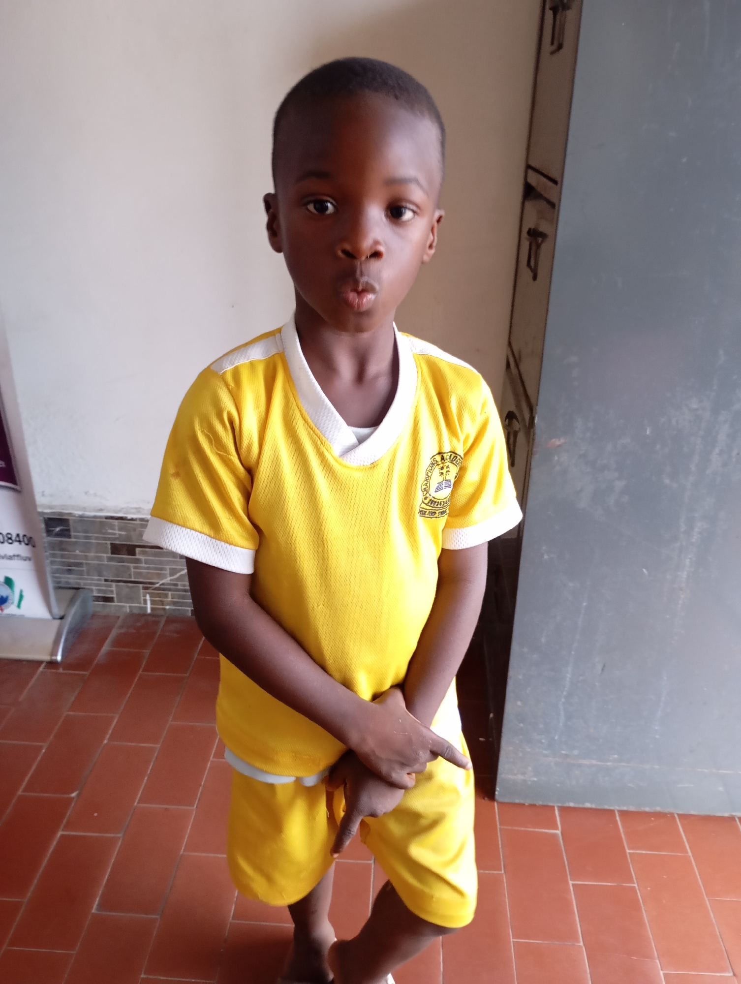 A young boy stands in front of a pale wall and a large metal cabinet. He is wearing a bright yellow t-shirt with white trim and matching yellow shorts. On his left chest is a school emblem with text that appears to read "CHAMPIONS ACADEMY". His hands are clasped in front of him, and his right index finger is extended, pointing slightly downwards. He has a curious or playful expression, with his lips pursed. The floor is tiled in a reddish-brown color. The lighting suggests it is daytime. To the left of the boy, a white stand with some text and graphics is partially visible. The scene appears to be outdoors or in a well-lit indoor space, possibly a schoolyard or hallway in Jos, Nigeria.