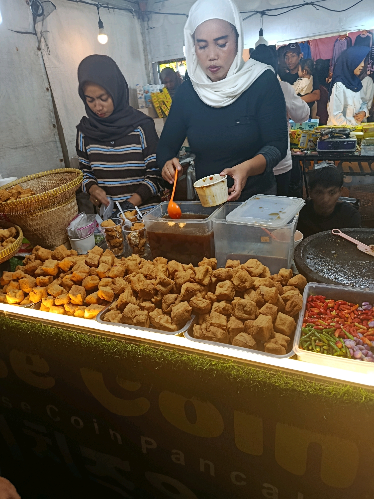 The image shows a street food stall in Balaraja, Indonesia, likely at a night market. In the foreground, a large quantity of fried tofu is displayed on a metal tray, illuminated by bright lights. To the right of the tofu, a plastic container holds a mix of red chilies, green chilies, and red onions. Behind the tofu, two women are visible. One, wearing a black and brown striped shirt and a dark hijab, is tending to the stall. The other, in the center and more prominent, wears a black long-sleeved shirt and a white hijab, holding a small white cup with an orange utensil. In the background, several people are gathered, some holding children, and various products are displayed on shelves. The stall is set up under a white canopy, with an overhead light illuminating the scene. The lower part of the image shows a dark counter with a large yellow logo that appears to spell out "SECOIN PANCA". The overall atmosphere suggests a bustling commercial environment.