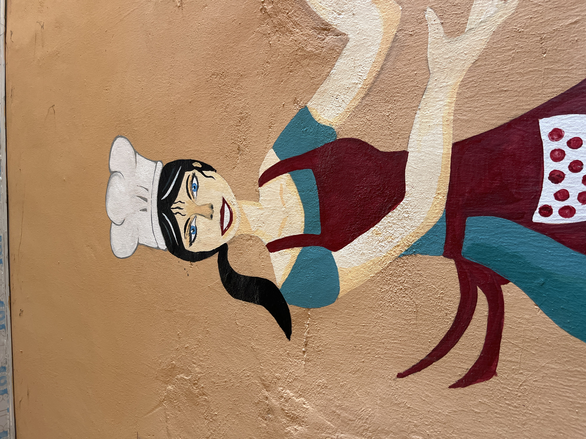 This is a photograph of a mural painted on a textured, light brown wall. The mural depicts a cartoonish figure of a woman dressed as a chef. She has black hair pulled back in a ponytail, a white chef's hat, bright blue eyes, and a wide smile with red lips. She is wearing a blue dress with red straps and an apron with a white top and red polka dots. Her arms and legs are outstretched, giving the impression of movement or a playful pose. The art style is simplistic and bold, with thick black outlines and flat areas of color. The background is the plain wall itself, suggesting an outdoor or public setting. The overall mood of the artwork is cheerful and lively. There is no visible text in the image.