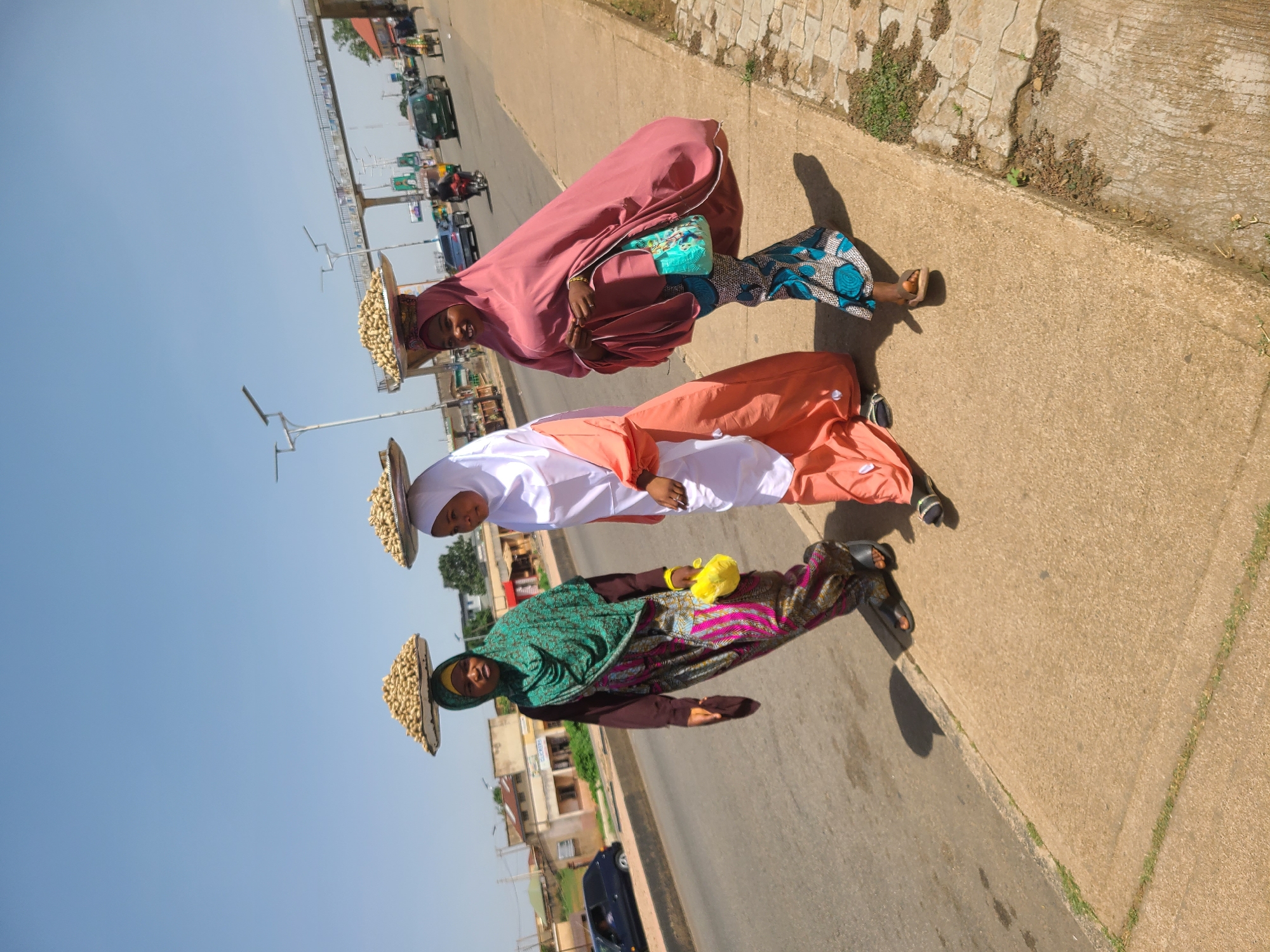 Three women walk along a city sidewalk. They are dressed in traditional clothing, including headscarves. Each woman carries a large, flat basket on her head filled with what appears to be peanuts. The sidewalk is beside a street with cars and lampposts, and buildings can be seen in the background.