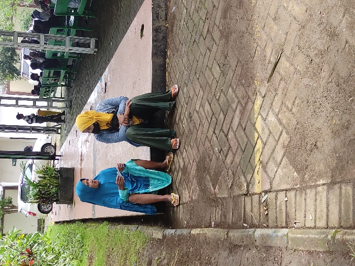 This image, captured in Kepanjen, Indonesia, presents an outdoor scene featuring several individuals in what appears to be a public waiting area during the daytime.

In the foreground, two women are seated on the ground beside a paved walkway. The woman on the left wears a blue hijab and a blue patterned dress, holding an object in her hands. Next to her, another woman is dressed in a yellow hijab, a light blue long-sleeved top, and a dark green long skirt. She wears a face mask, has her arms crossed, and is looking downwards, suggesting a state of rest or contemplation. Both women are wearing sandals.

Behind them, the scene extends to a sheltered area with several people, mostly men, sitting on green benches, indicative of a waiting zone. A white car is partially visible parked further in the background. The ground is a mix of patterned paving bricks and an unpaved, dirt area to the right. A low concrete curb separates the walkway from a strip of green grass and small plants.

The bright, natural lighting suggests it is daytime with clear weather. The overall impression is one of people waiting or resting in a public outdoor space, possibly outside a local facility or during a public gathering. There is no visible text in the image.