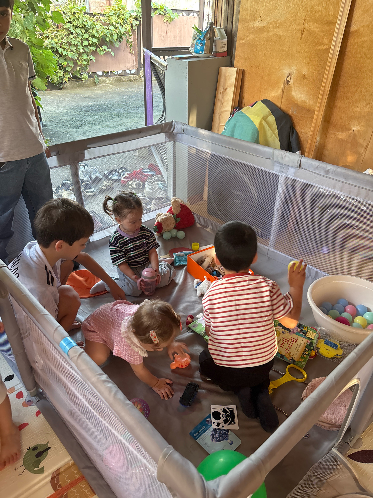 In a domestic setting, possibly a garage or covered outdoor area, a playpen contains four young children. A boy in a white t-shirt with black lettering is on the left, facing inwards. A girl with a ponytail, wearing a striped t-shirt, is sitting on the right, holding a pink bottle. A baby in a pink romper is crawling in the center. Another boy in a red and white striped shirt is on the far right, facing away from the camera, holding a yellow ball. The playpen is filled with various toys, including a tub of colorful balls, small cars, and stuffed animals. Outside the playpen, shoes are visible on the ground. The environment appears to be slightly cluttered, with wooden walls and foliage visible in the background.
