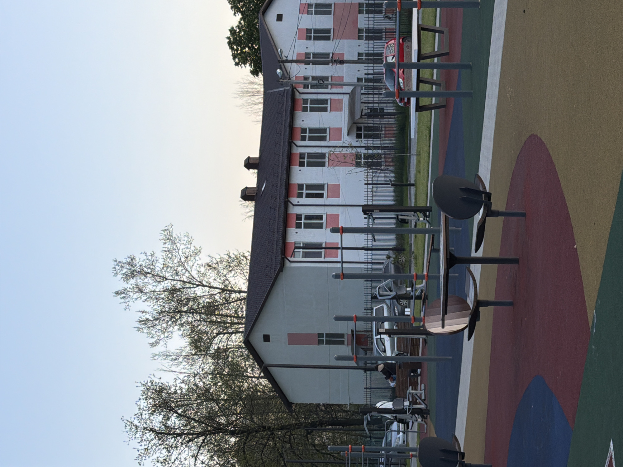This image captures an outdoor sports ground or workout area situated adjacent to a residential building in Beryozovy, Russia. The foreground features a vibrant, multi-colored padded surface, adorned with geometric patterns in shades of red, blue, green, yellow, and brown. Spread across this surface are various pieces of outdoor exercise equipment, including parallel bars, pull-up stations, and stationary fitness machines, many with dark metal frames and some featuring wooden accents.
In the background, a multi-story residential building with light-colored walls and distinct pink or red accents around its windows stands prominently. It has a dark, pitched roof. A metal fence separates the sports facility from the area around the building, where several cars are parked, including a visible red vehicle. Trees with budding or sparse leaves are visible against a clear, pale blue sky, suggesting the time of day is either late afternoon or early evening. Power lines and poles are also present near the buildings.
One person is partially visible seated on a bench or equipment in the mid-ground, and another individual's legs are faintly seen further back. There is no overt activity taking place; the scene appears calm with individuals either resting or preparing to use the equipment. No text is visible in the image.